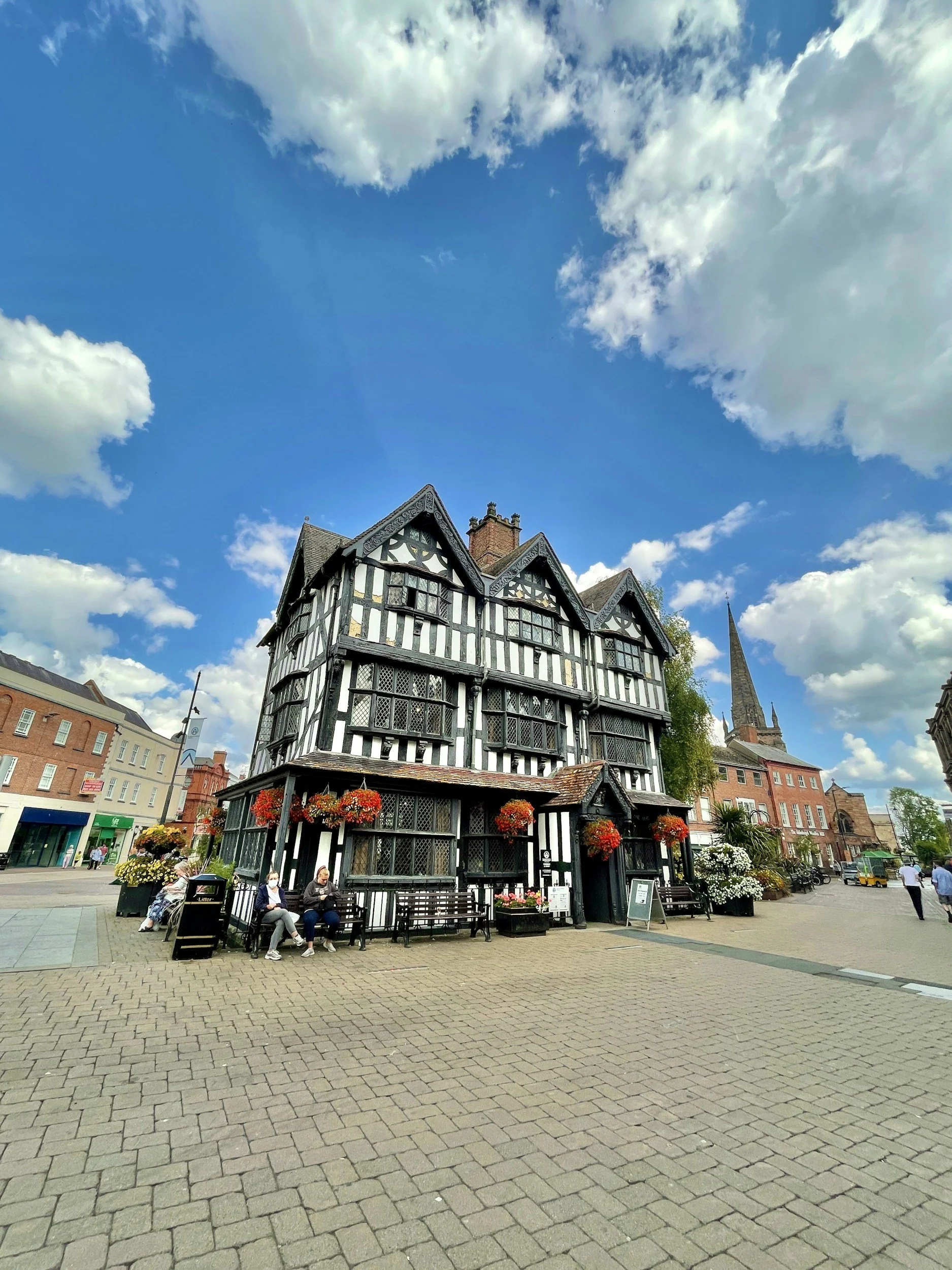 A historic black and white timber-framed building in a town square with benches, people sitting, and hanging flower baskets, under a blue sky with clouds.