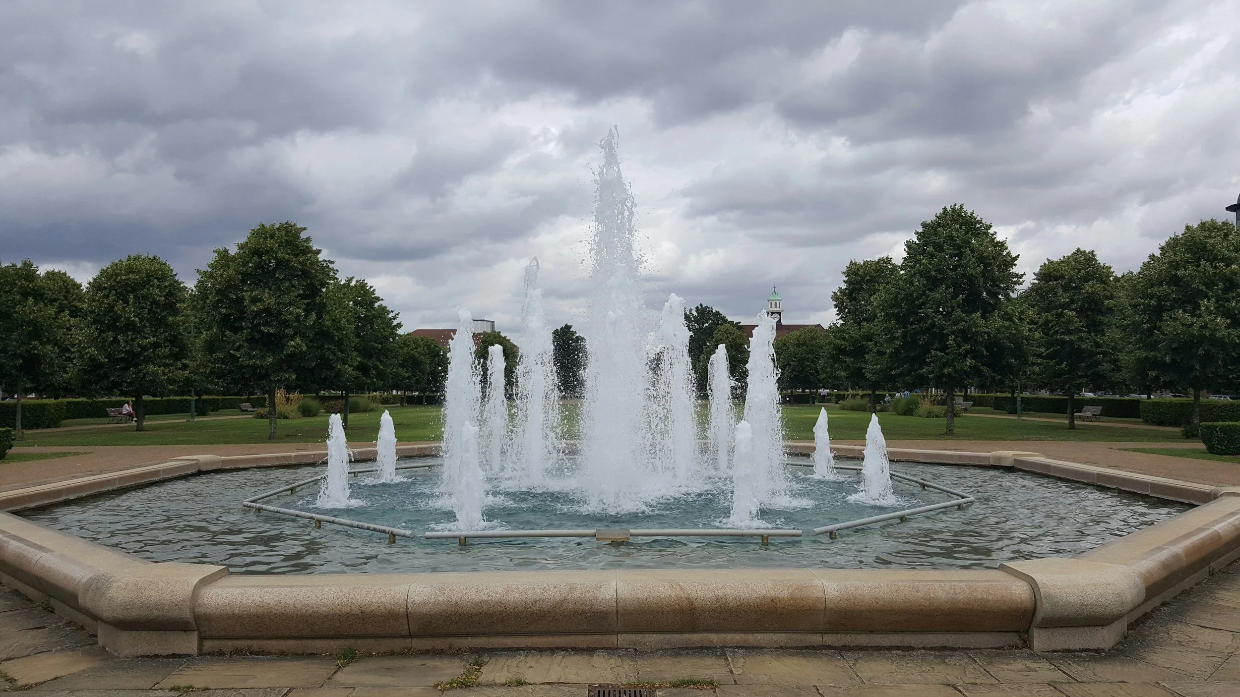 A fountain with multiple water jets in a park with trees and a cloudy sky.