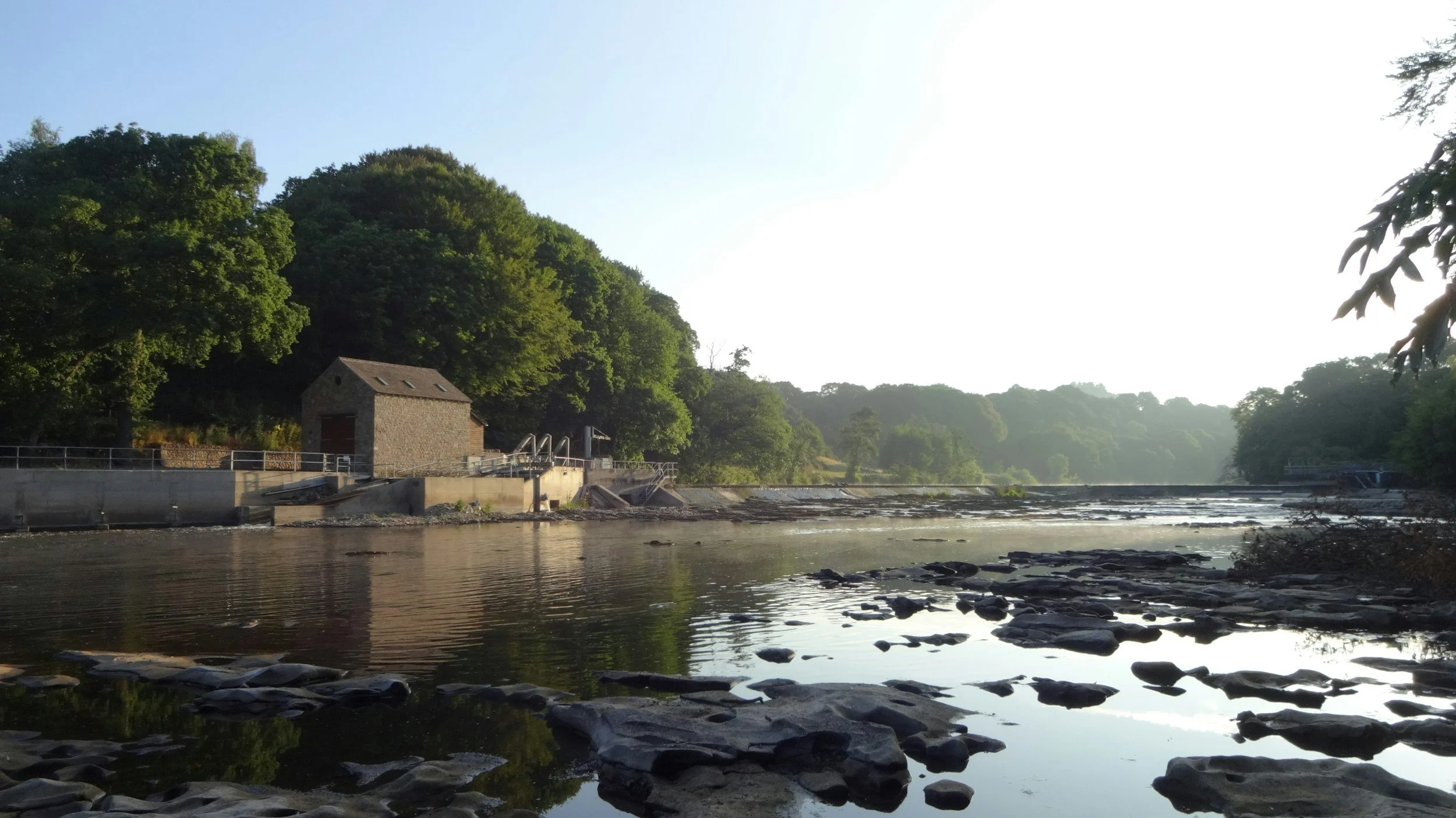 A river with rocks along the shore, trees on the opposite bank, and a small stone building near the water, under a clear sky.