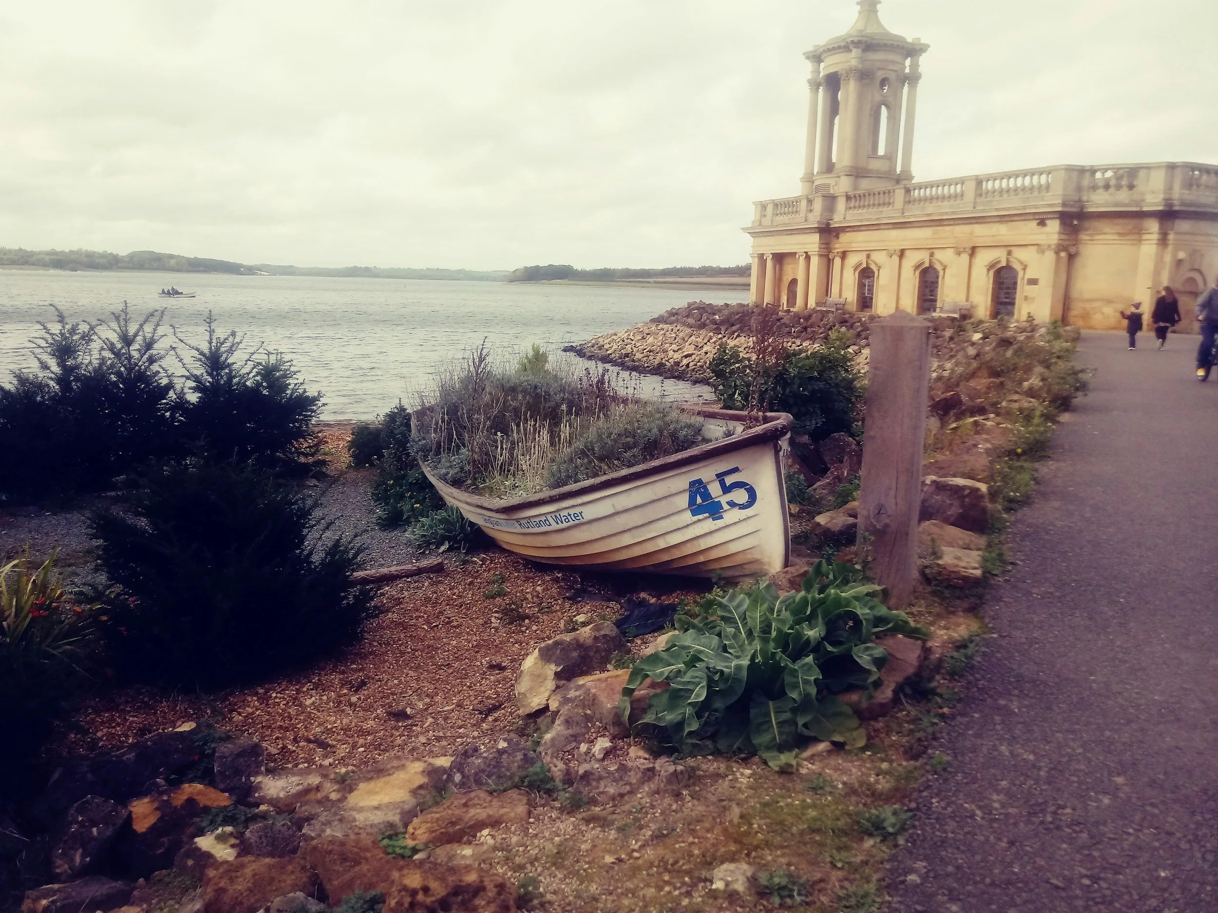 A boat on a large body of water with a historic building on the right and people walking along the shoreline.