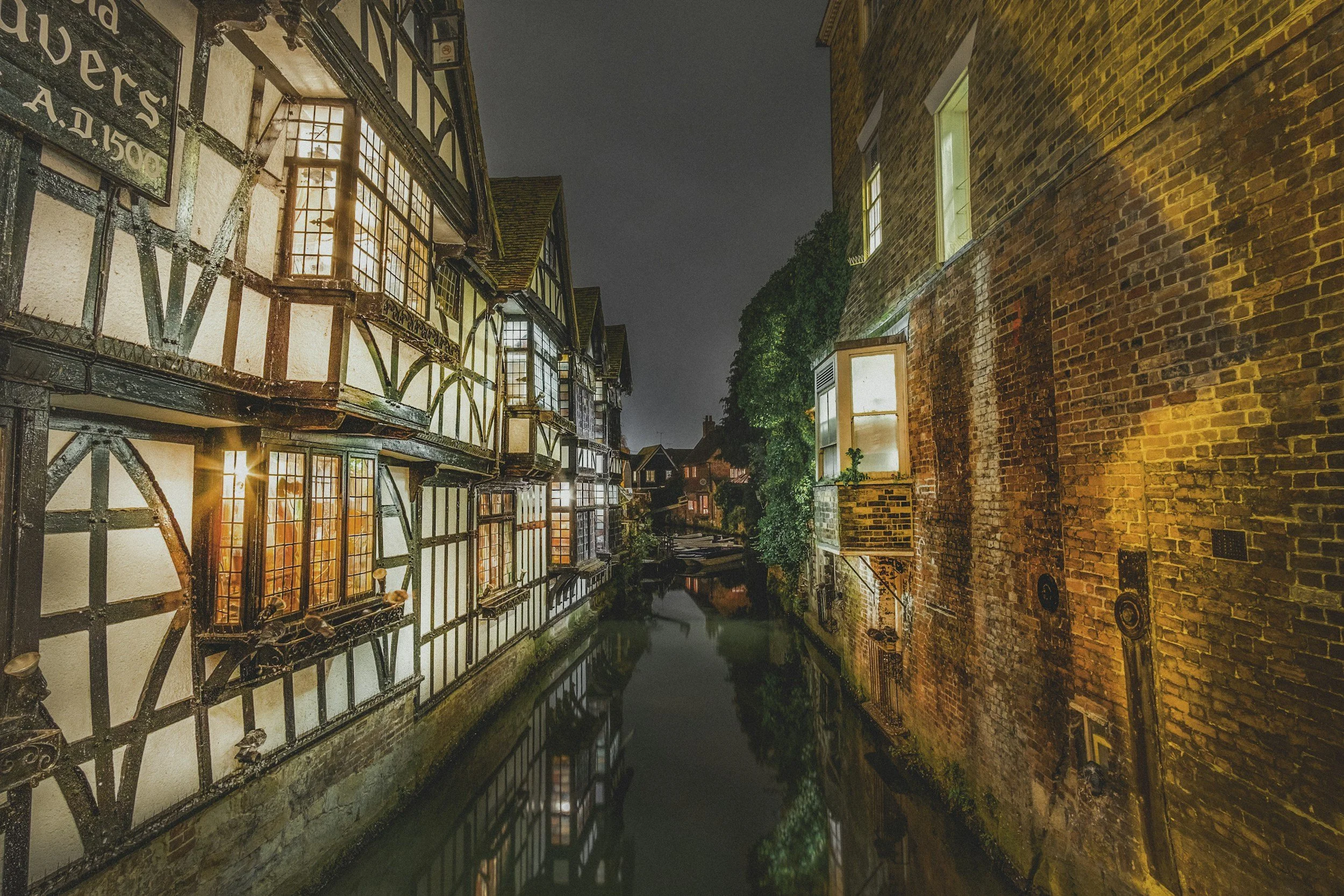 Nighttime view of a narrow canal flanked by historic half-timbered houses on the left and brick buildings on the right, with the canal's water reflecting the illuminated windows of the buildings.