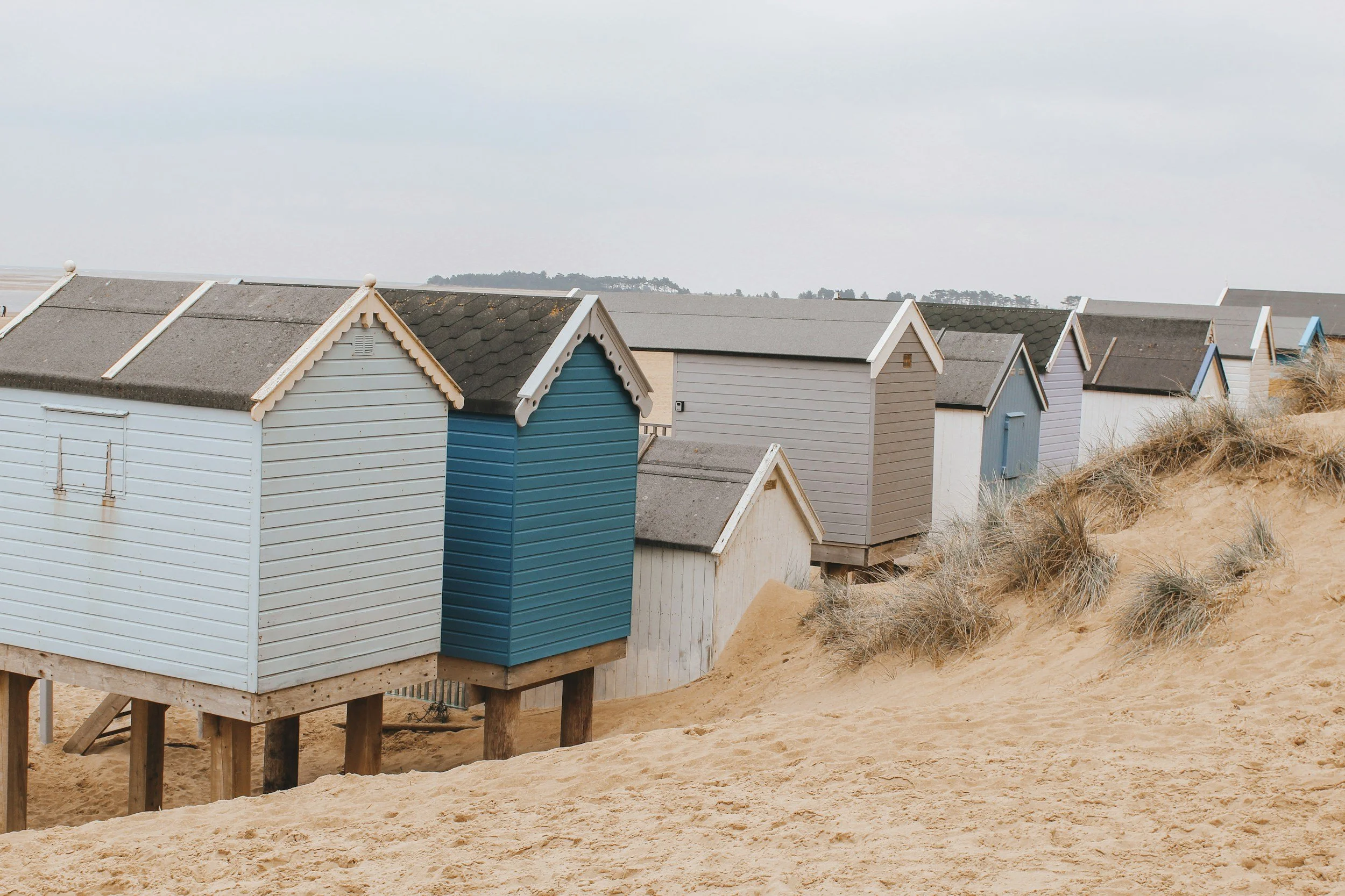 Colorful beach huts on stilts above sandy dunes with sparse grass, under a cloudy sky.