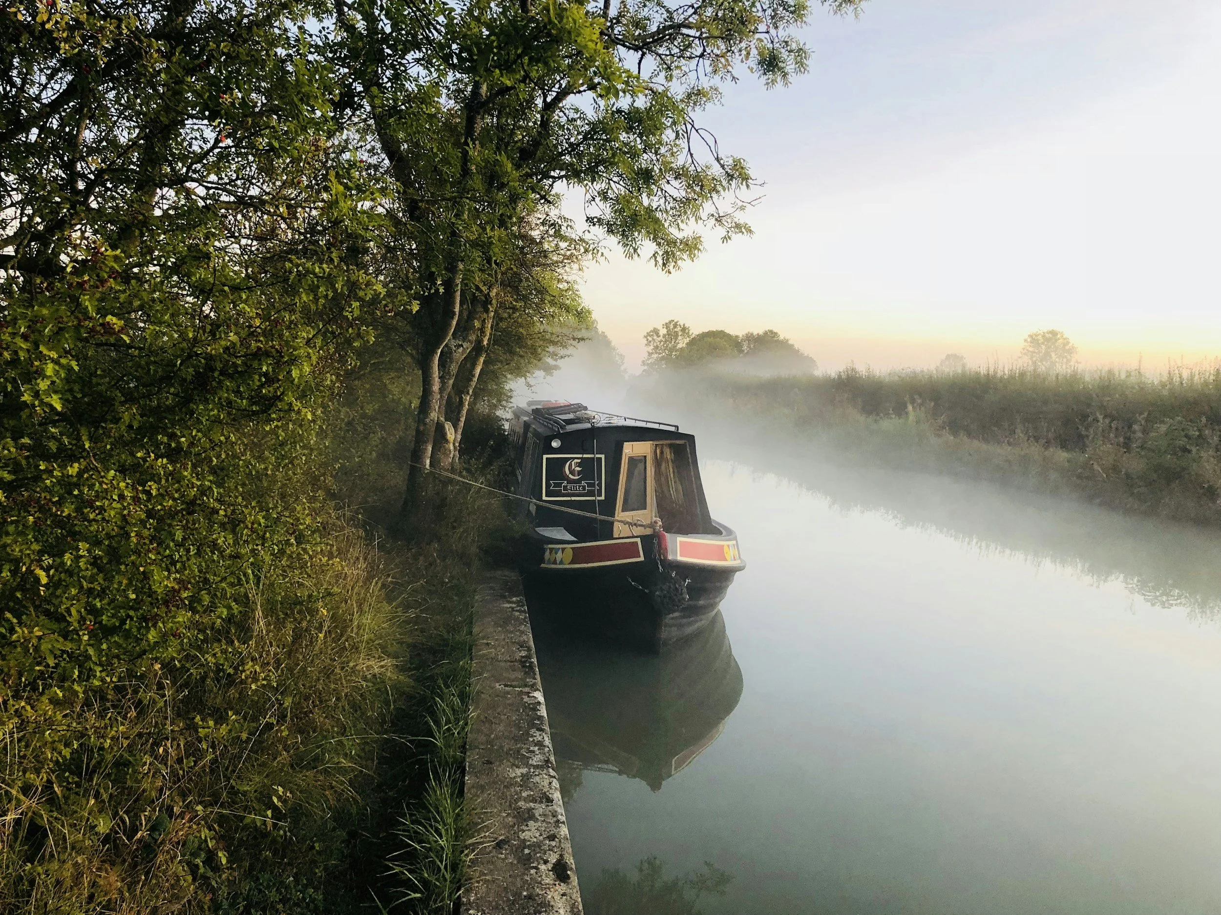 A narrowboat moored along a calm riverbank surrounded by lush green trees, with mist rising from the water and a soft sunrise or sunset in the background.