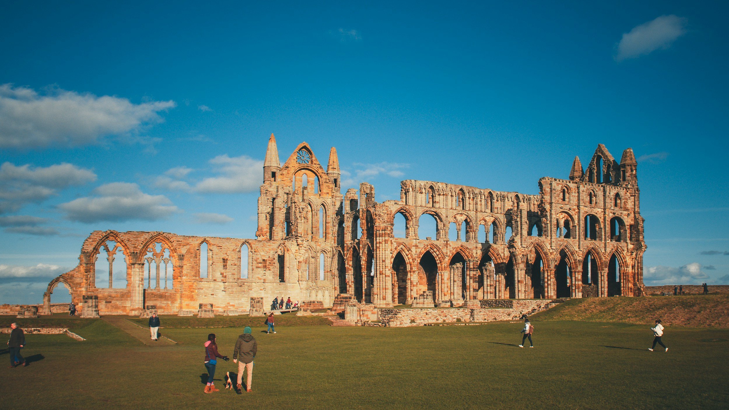 Ruin of an ancient stone church with Gothic arches and towers under a blue sky with clouds, with people walking and exploring the site.