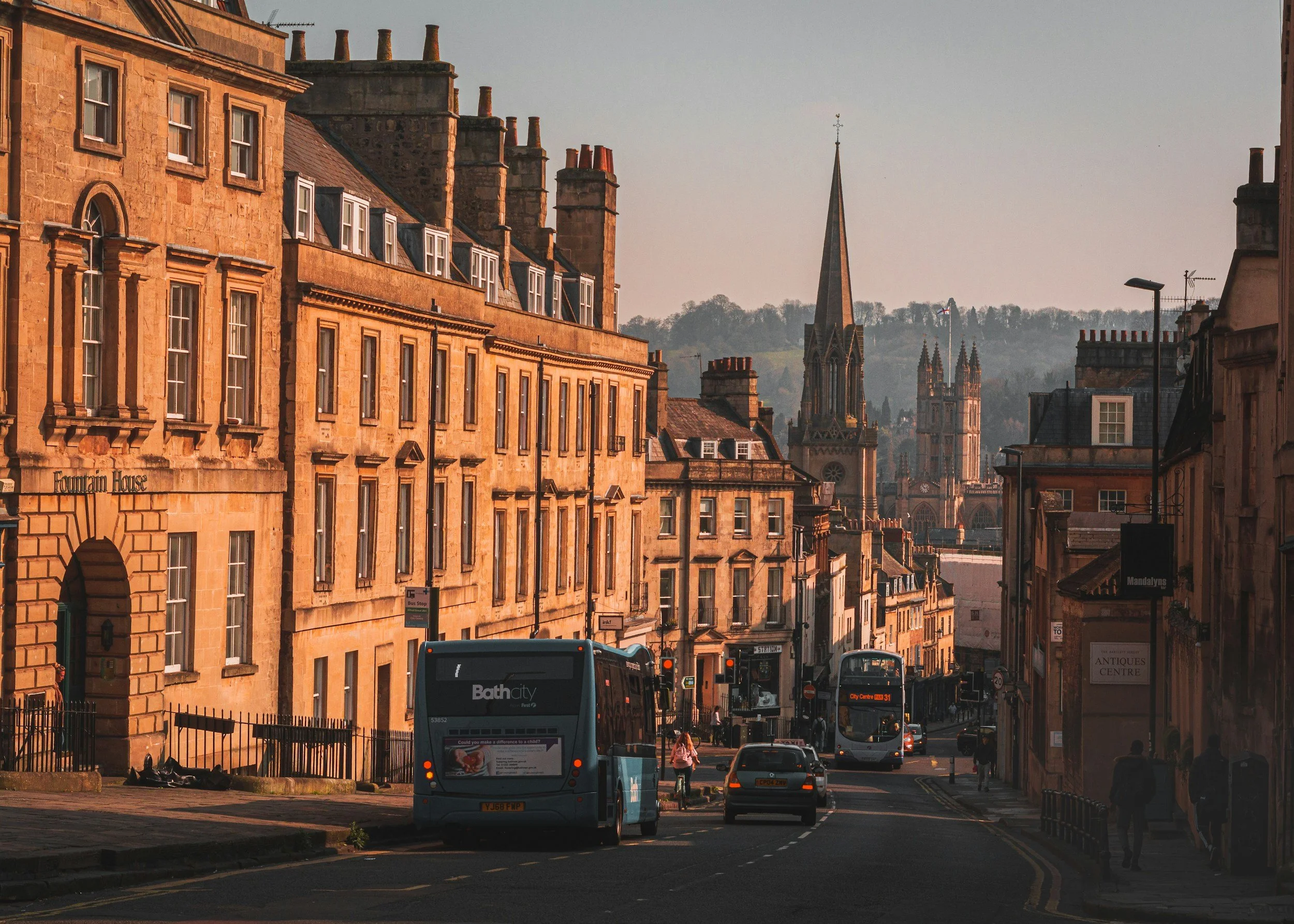 Street scene with historic brownstone buildings, a church with spires in the background, and buses on the road during sunset.