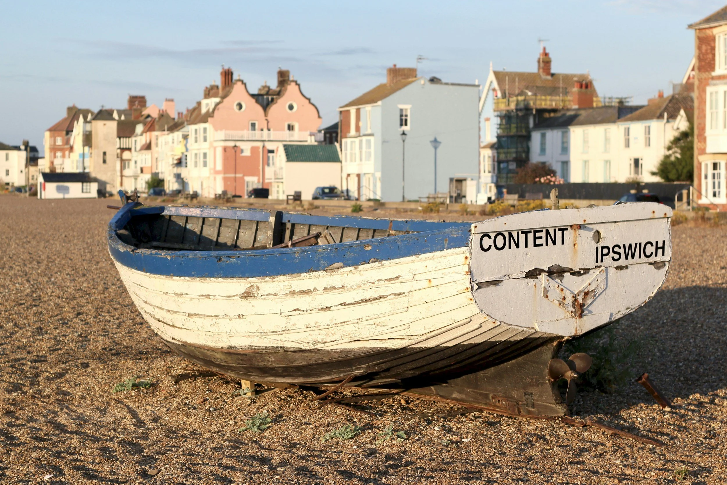 An old white boat with chipped paint and rust, labeled "CONTENT IPSWICH," resting on a pebbled beach with colorful houses in the background.