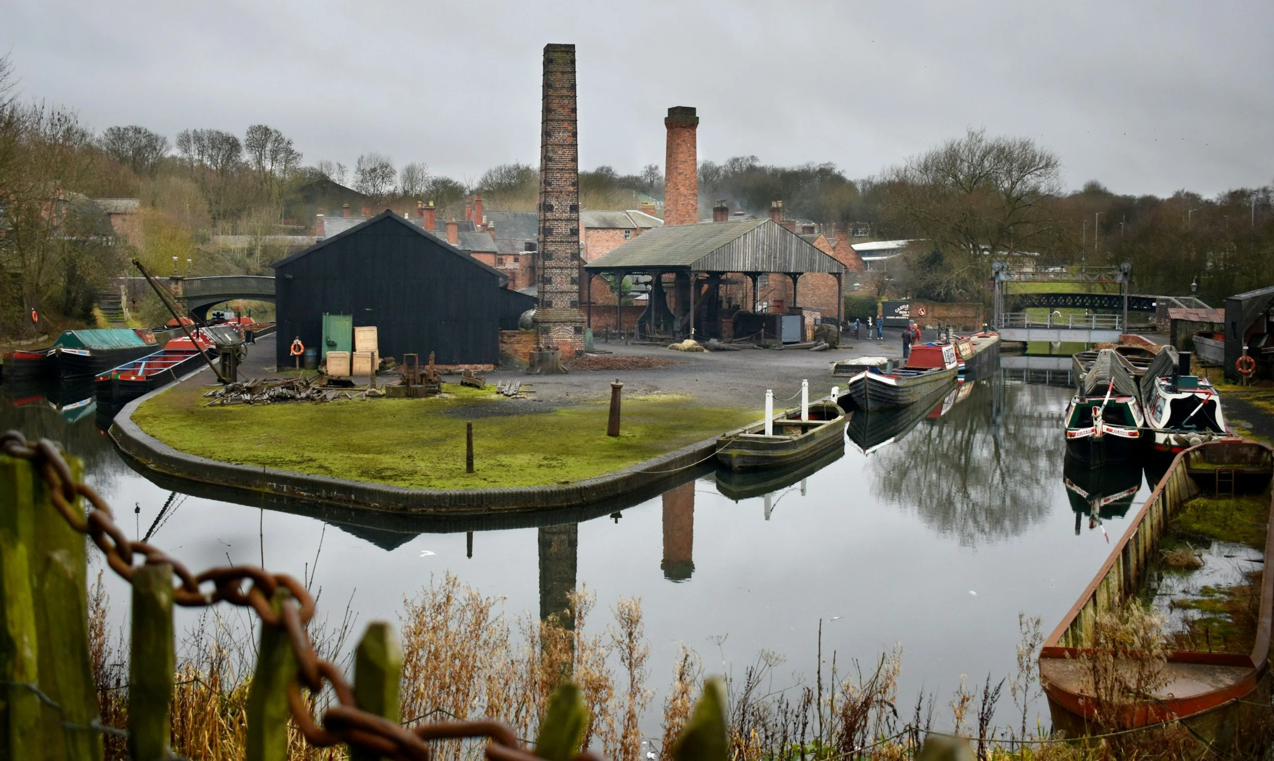 A historic canal with moored boats, surrounded by industrial buildings with tall chimneys, trees, and a bridge in the background, overcast sky.
