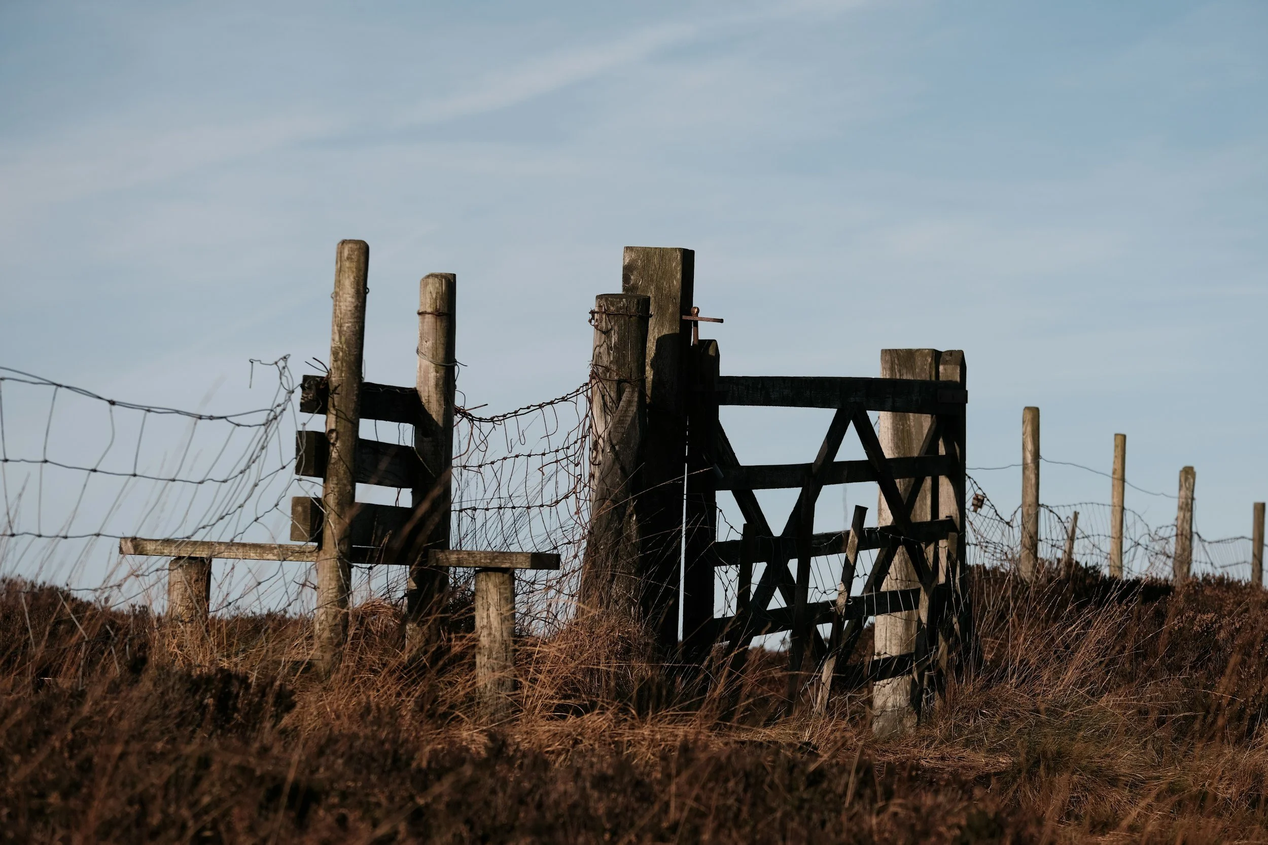 A weathered wooden fence with barbed wire, part of a broken or abandoned structure, set against a clear blue sky with some clouds.