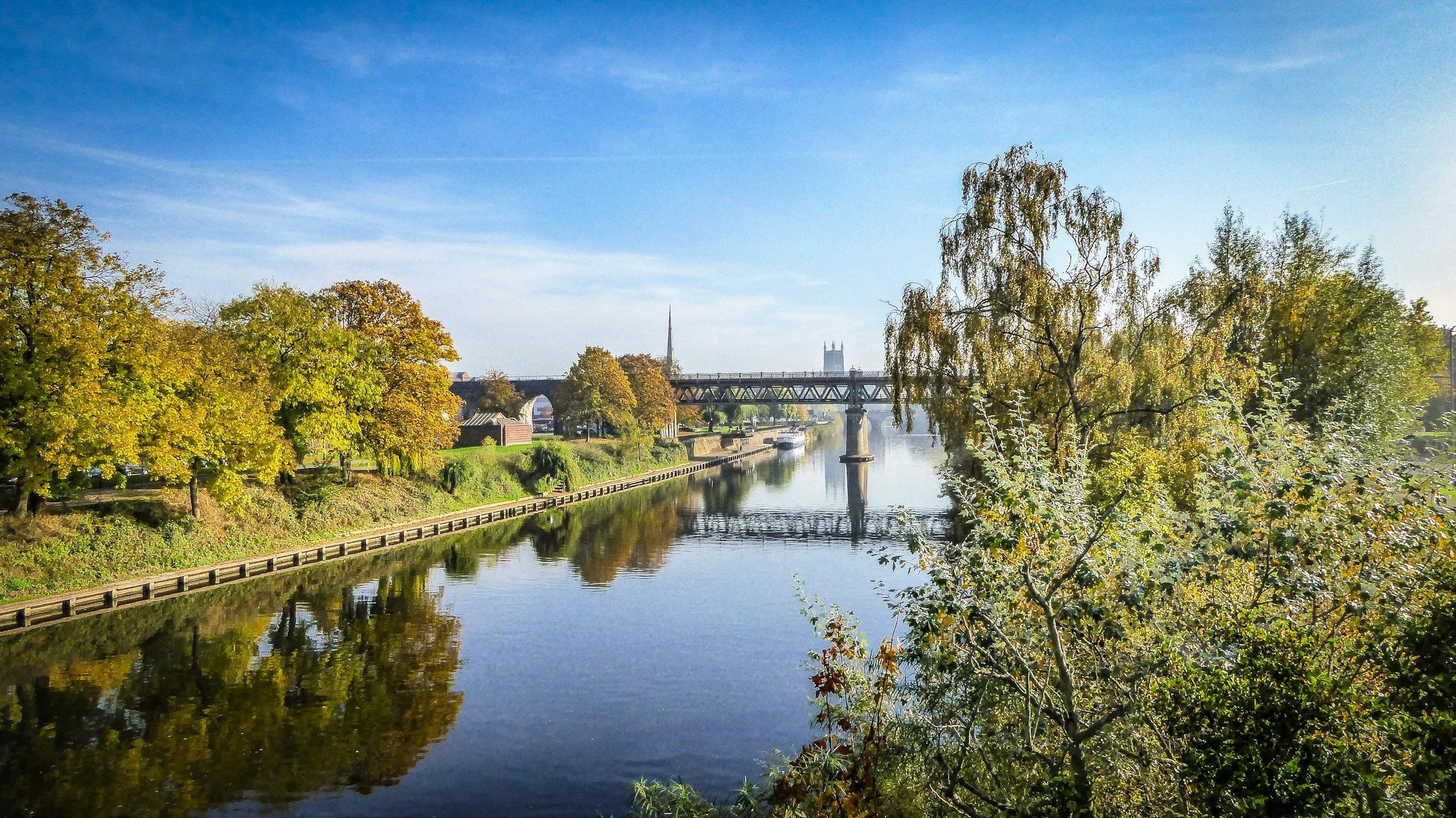 A calm river flowing through a city, with trees lining the banks and a bridge crossing over it. The sky is blue with some clouds.