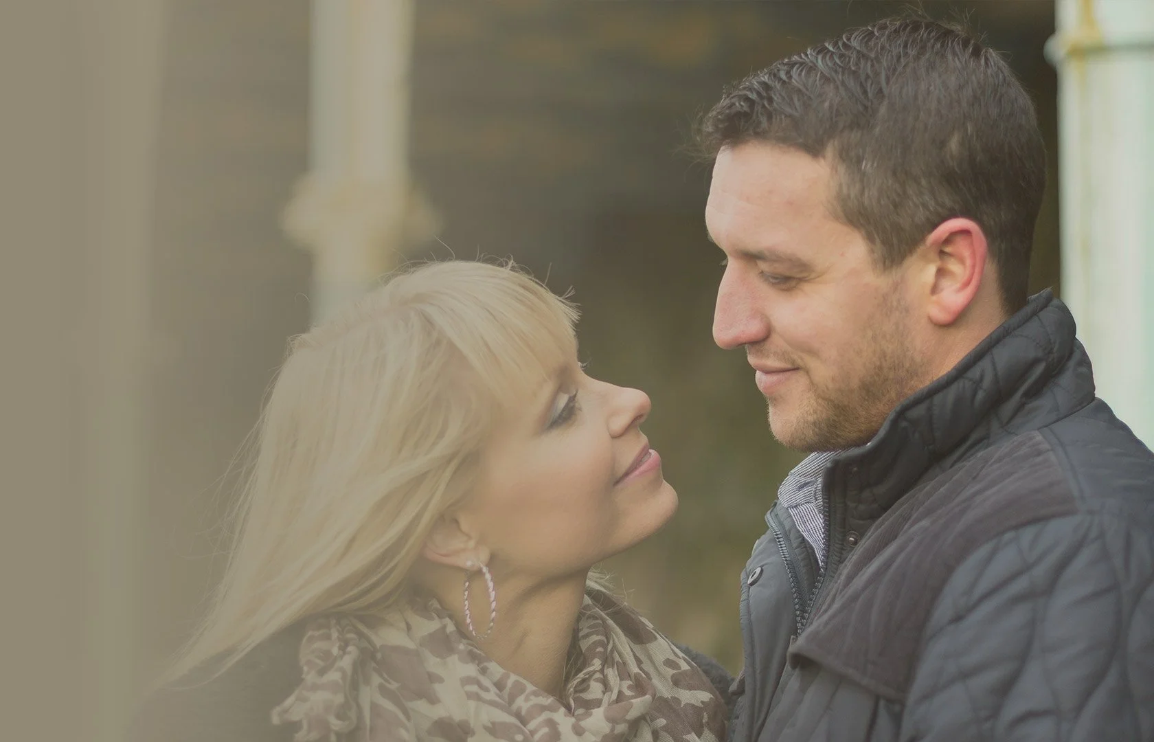 A man and woman are standing close together outdoors, gazing into each other's eyes with affection. The woman is smiling slightly, and the man has a gentle expression. The background appears to be a natural setting.