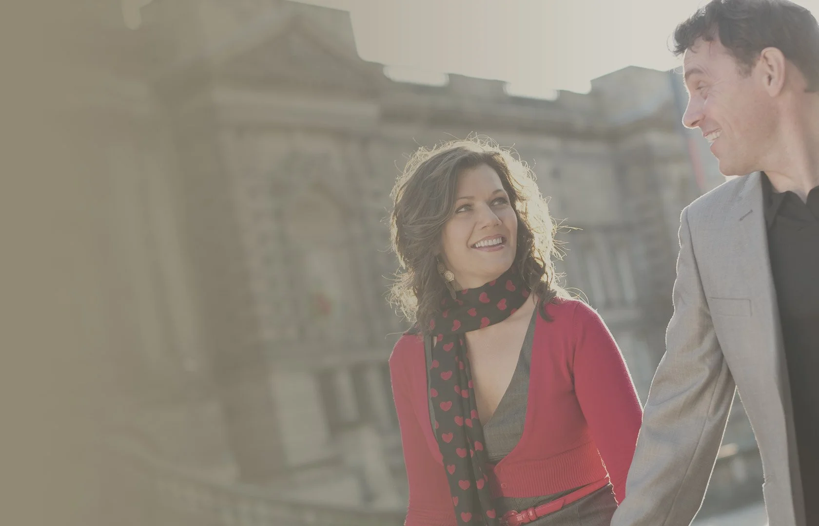 A woman and a man smiling and looking at each other while walking outdoors in daytime with old buildings in the background.