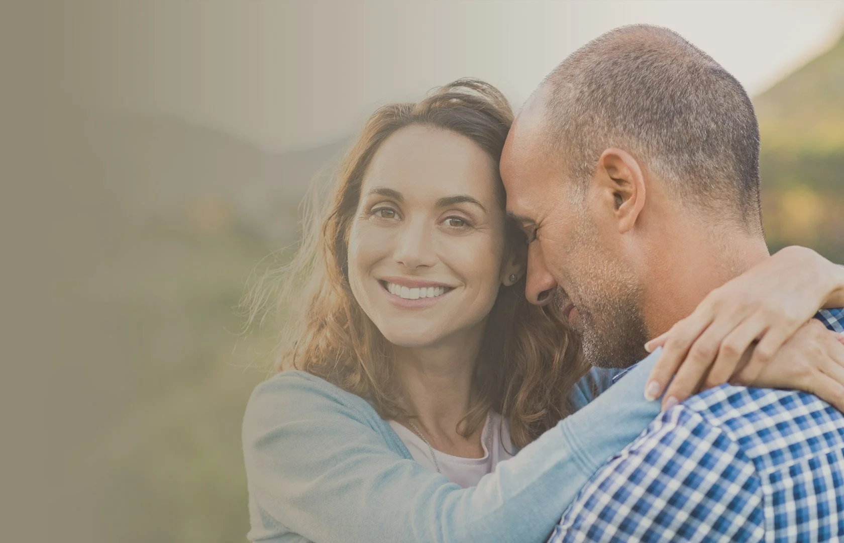 A smiling woman with brown hair embracing a man with a beard and short hair outdoors.