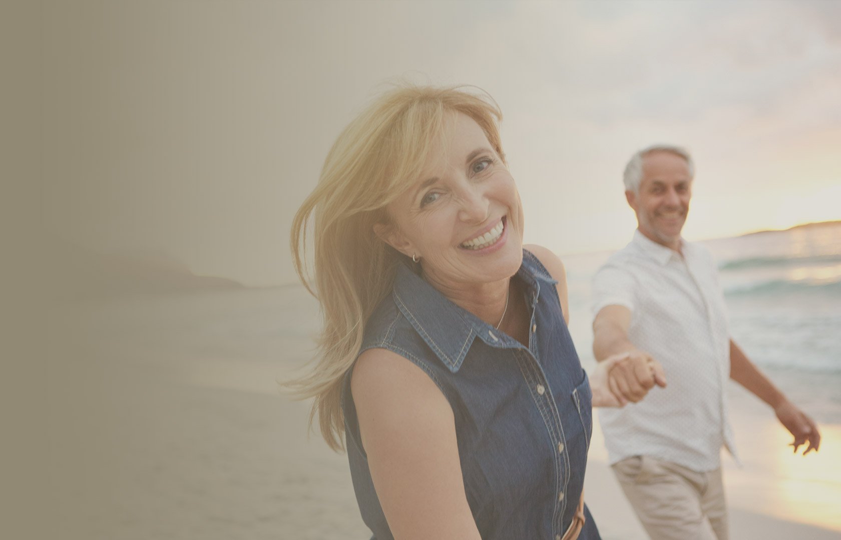 A smiling woman with blonde hair and a sleeveless denim shirt holding hands with a man with gray hair and a white shirt at the beach during sunset.