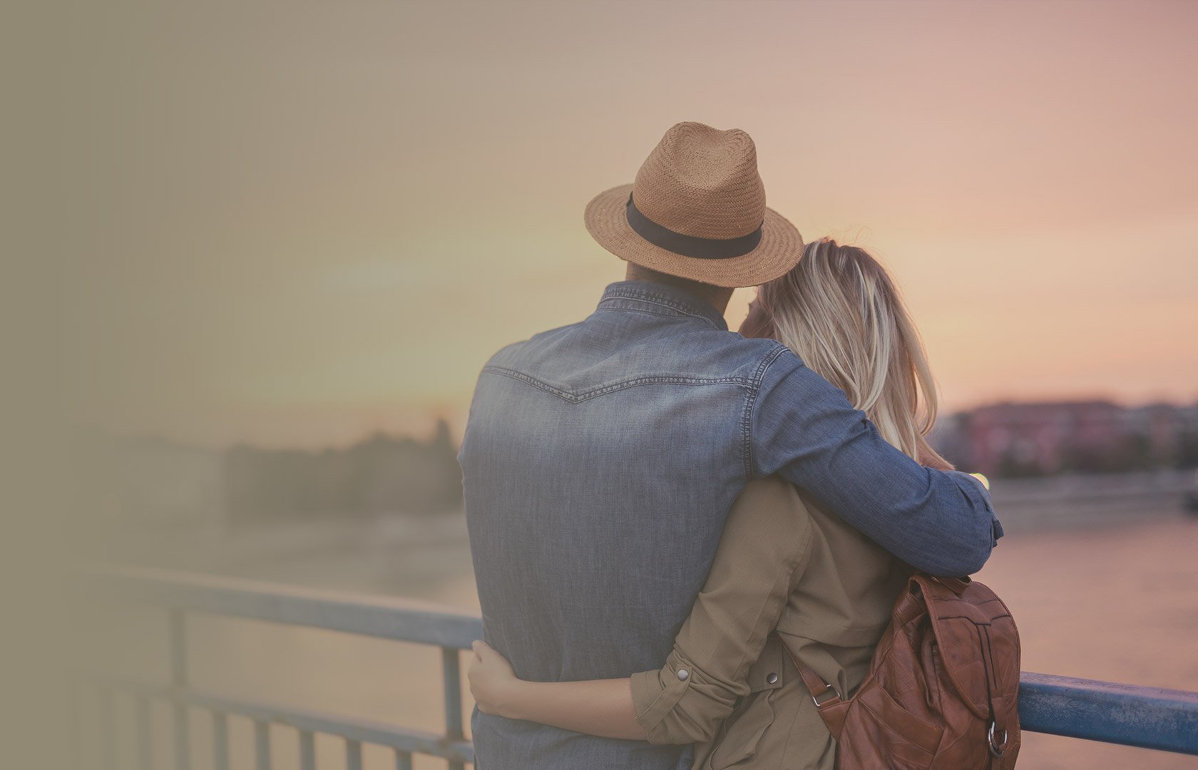 A couple is hugging on a bridge during sunset, overlooking a river and cityscape in the distance.