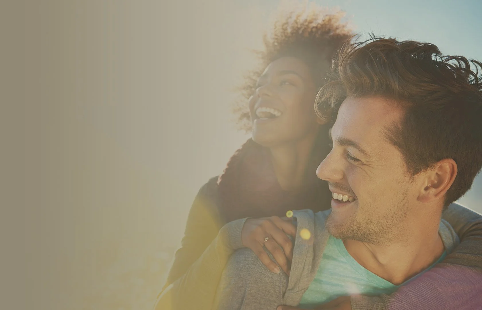 A happy young couple smiling outdoors in sunlight, with the woman showing her hand with a ring.