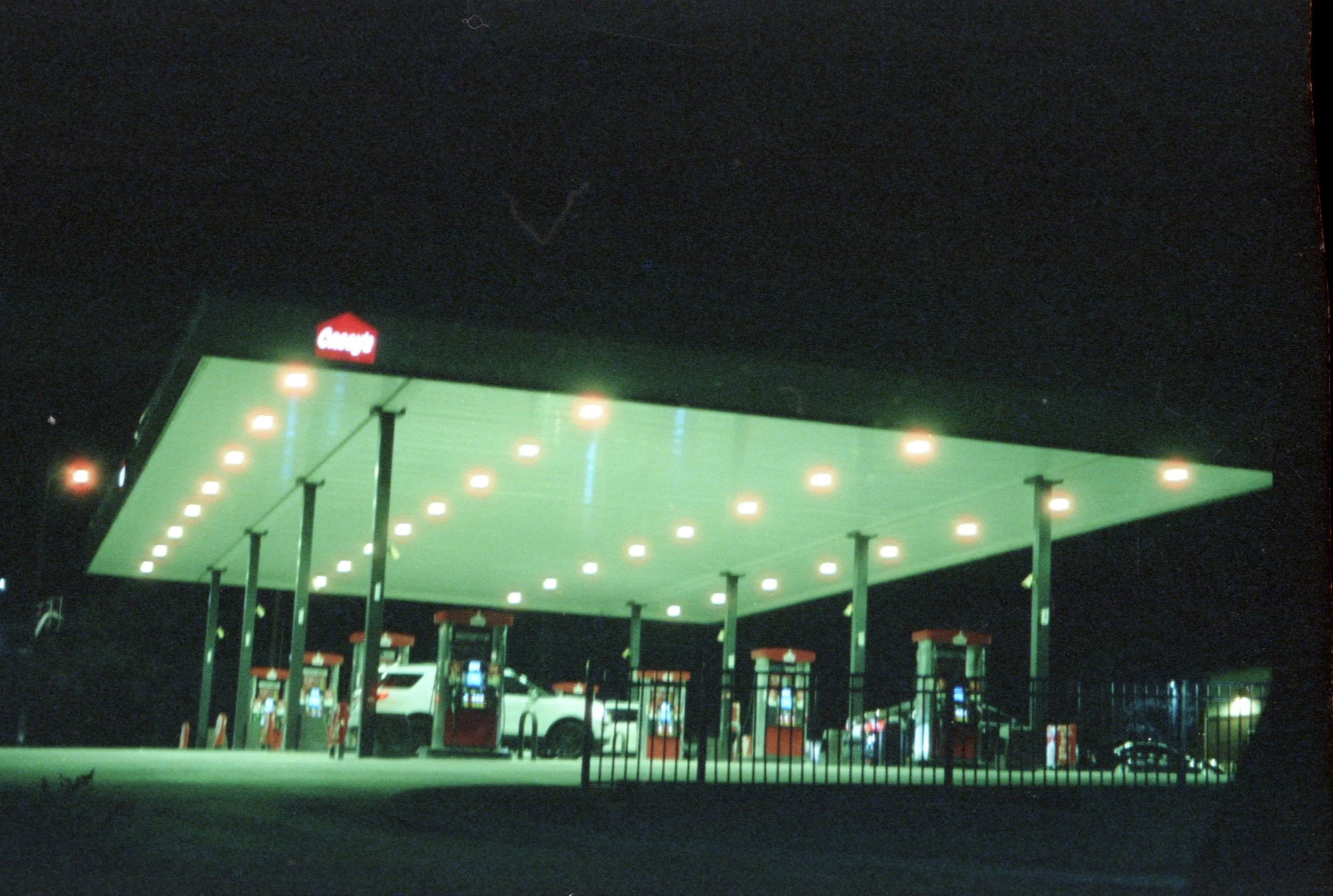Nighttime view of a gas station with green roofing and string lights, a red Canopy sign, and multiple fuel pumps.