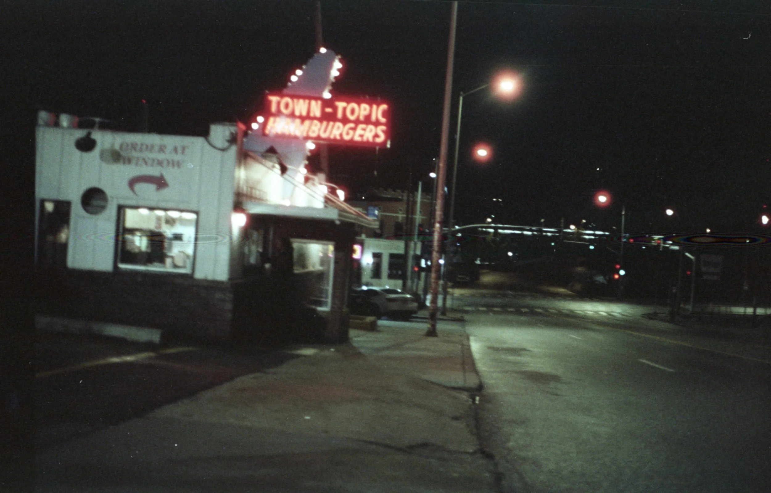 Nighttime view of a parking lot with a white food truck and illuminated sign that reads 'TOWN-TOPIC HAMBURGERS' in red lights. Several streetlights and parked cars are visible.