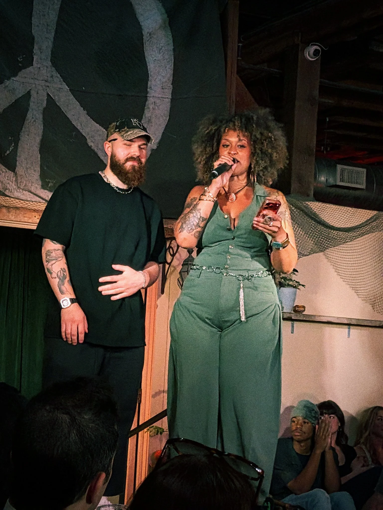 Two women standing on a stage with a dark background, one woman is speaking into a microphone and the other has her eyes closed with a slight smile. Several audience members are seated and watching.