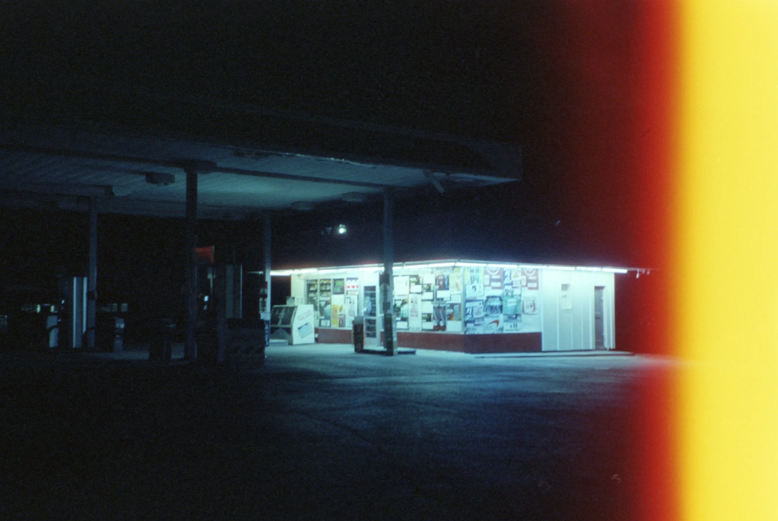 A dark outdoor scene of a closed convenience store illuminated by bright lights inside, with a display of magazines and snacks outside, and a small structure with a roof and support poles in the foreground. The right side of the image has a color lig
