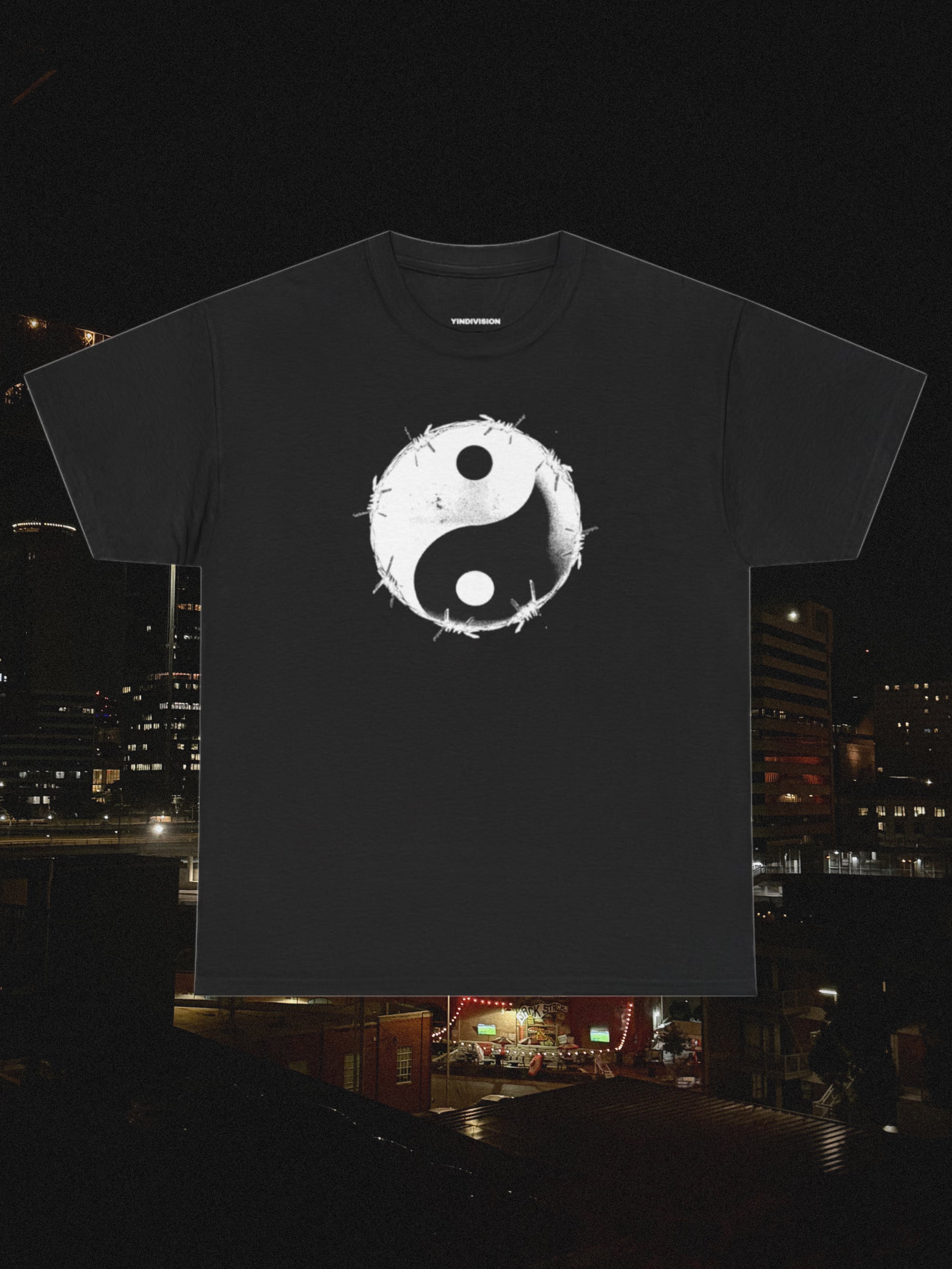 A black T-shirt with a white yin-yang symbol printed on the front, hanging against a city skyline at night.