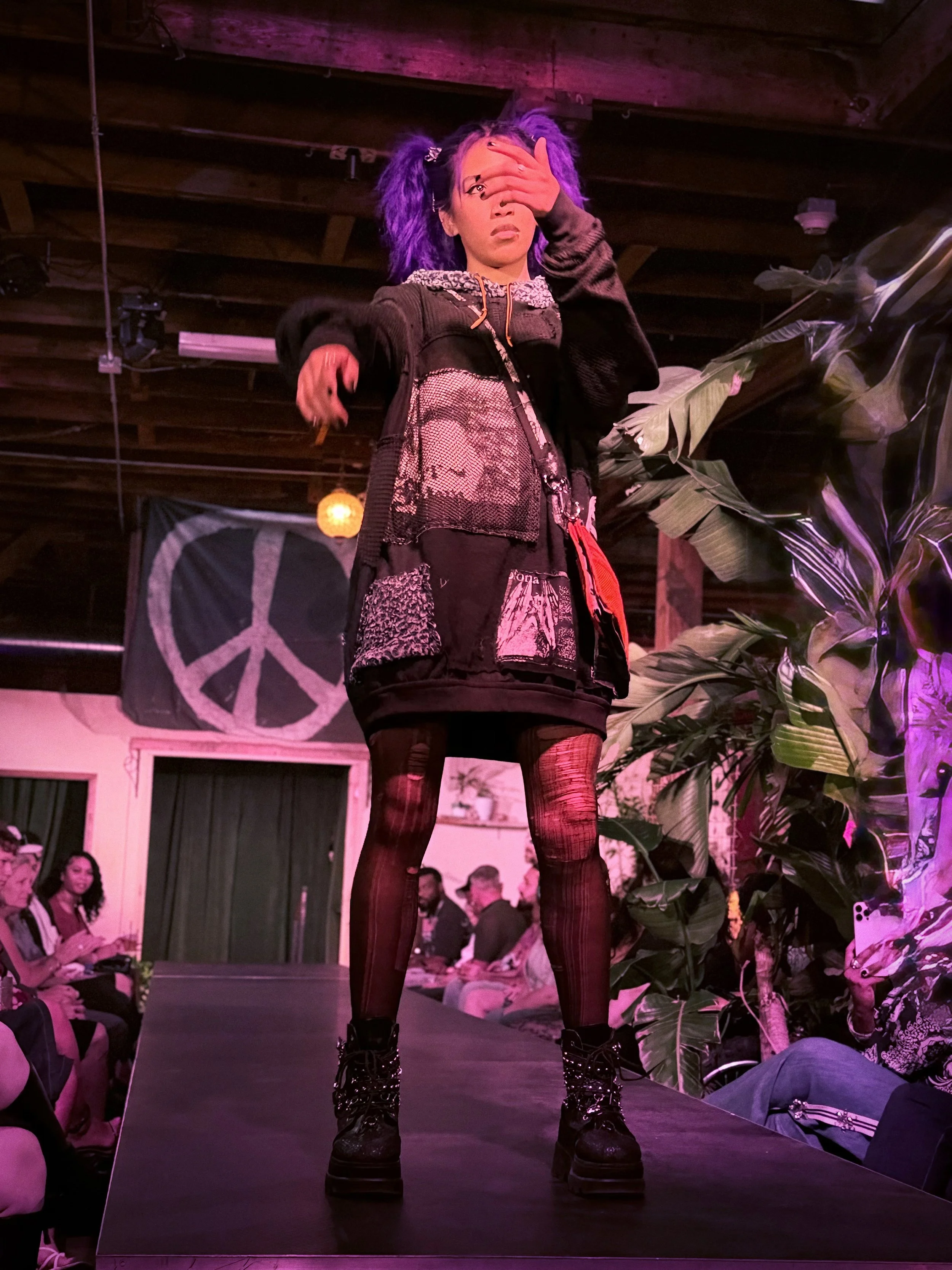 A person with purple hair, wearing a black patchwork outfit, fishnet stockings, and combat boots, walking down a runway during a fashion show with an audience in the background and a peace sign banner visible.