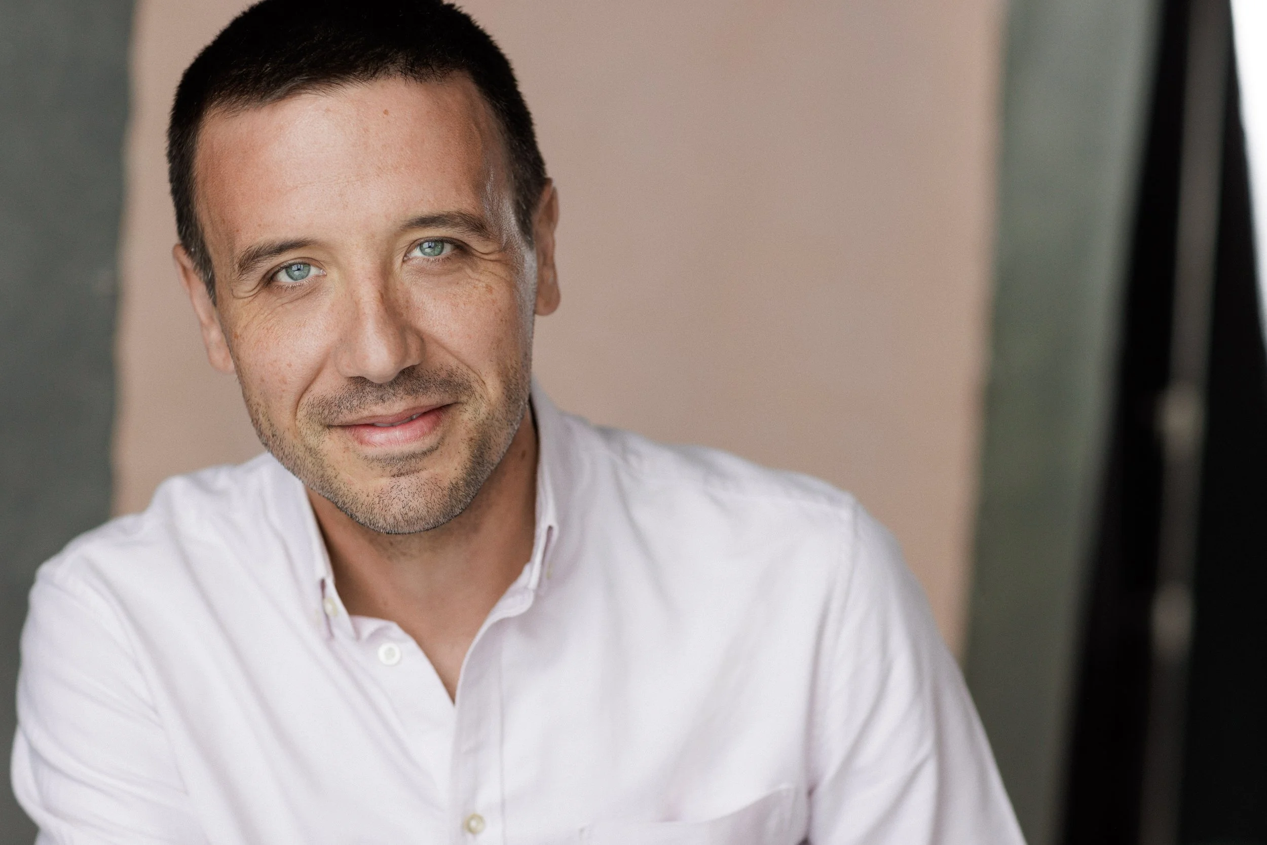 A smiling man with short dark hair, blue eyes, and light facial hair, wearing a white button-up shirt.