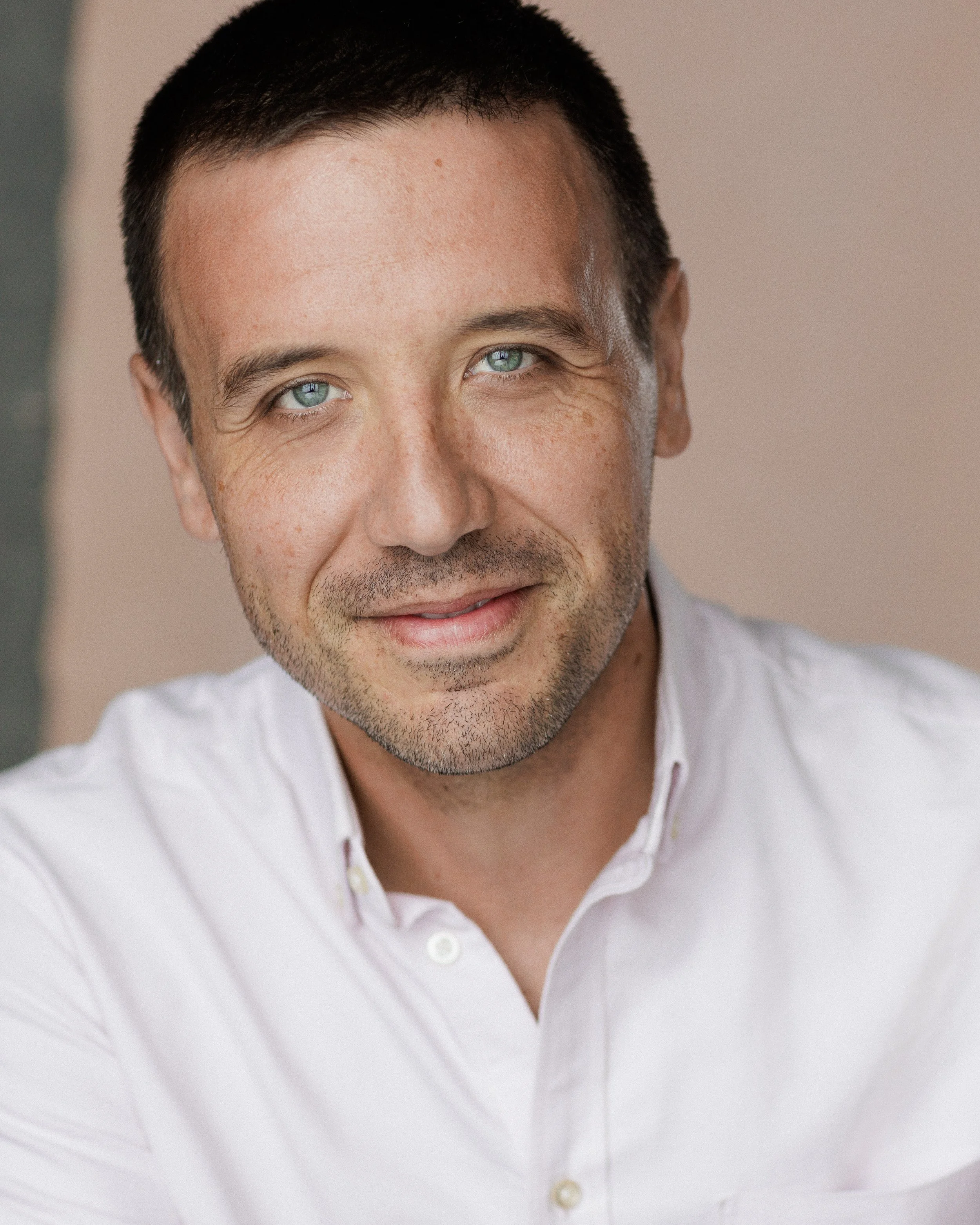 A close-up photo of a man with light skin, short dark hair, blue eyes, and slight facial hair, wearing a white button-up shirt, smiling with a natural expression, against a neutral background.