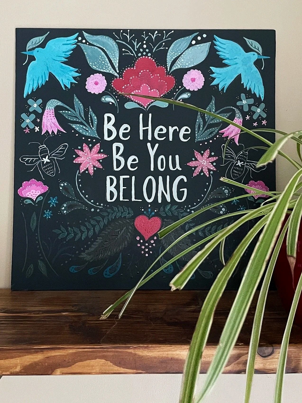 A decorative black chalkboard sign with colorful floral and bird designs and the message "Be Here Be You BELONG" written in white. A green houseplant partially covers the lower right corner.