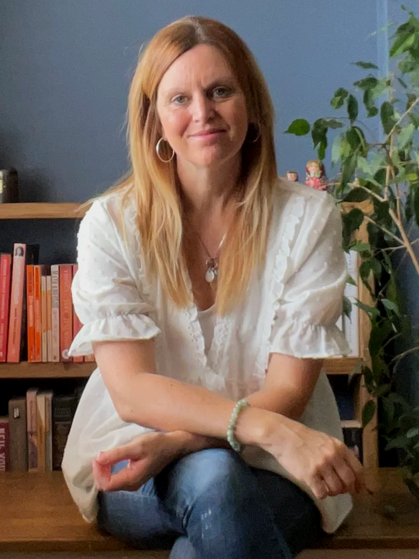 Smiling woman with long blonde hair sitting on a wooden bench in front of bookshelves, with a blue wall and a green plant in the background.