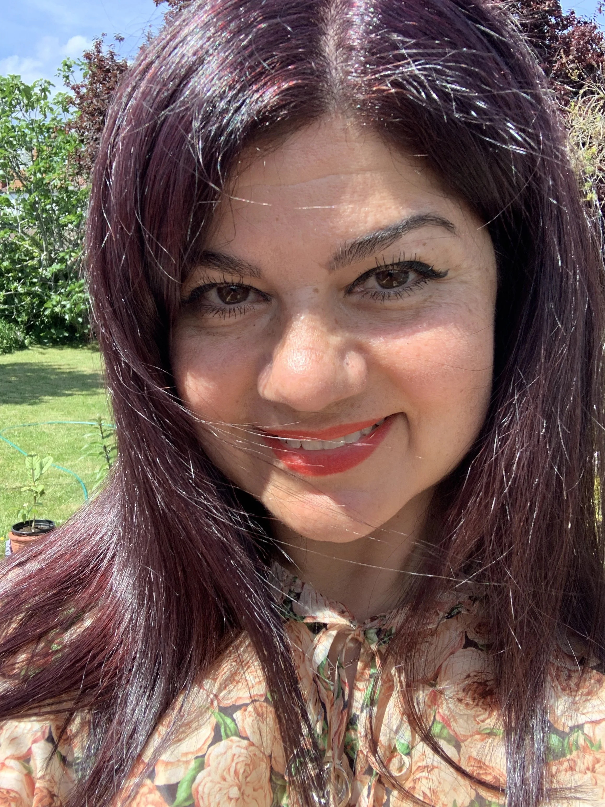 A woman with long, wavy, dark red hair smiling outdoors on a sunny day, wearing a floral-patterned beige top, with green trees and a blue sky in the background.