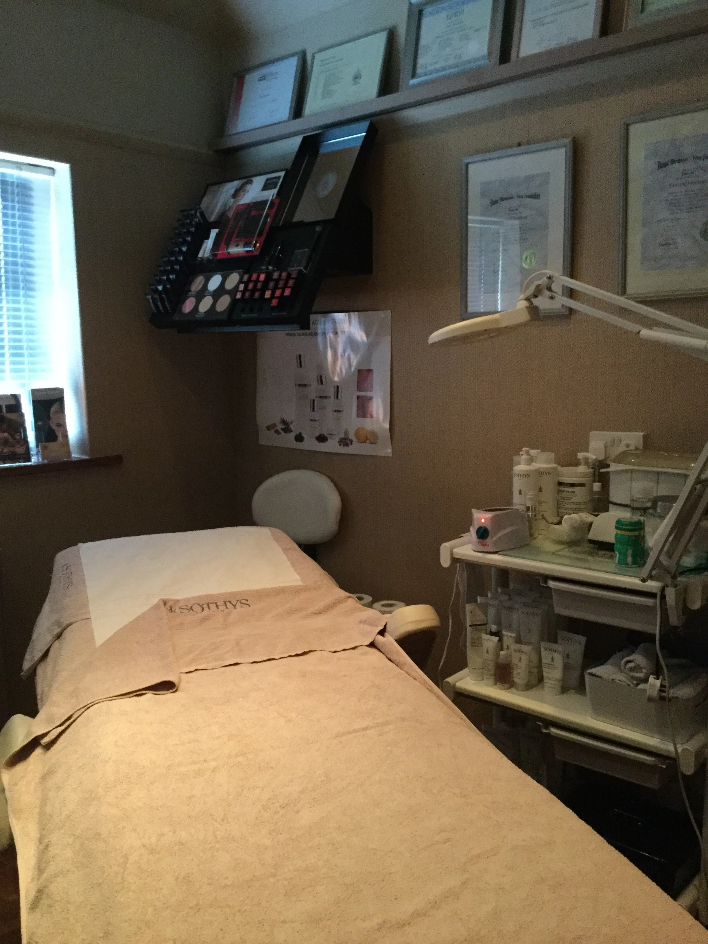 A medical treatment room with a bed covered in a beige towel, medical supplies and equipment on shelves, a lamp, framed certificates on the wall, and a window with blinds.