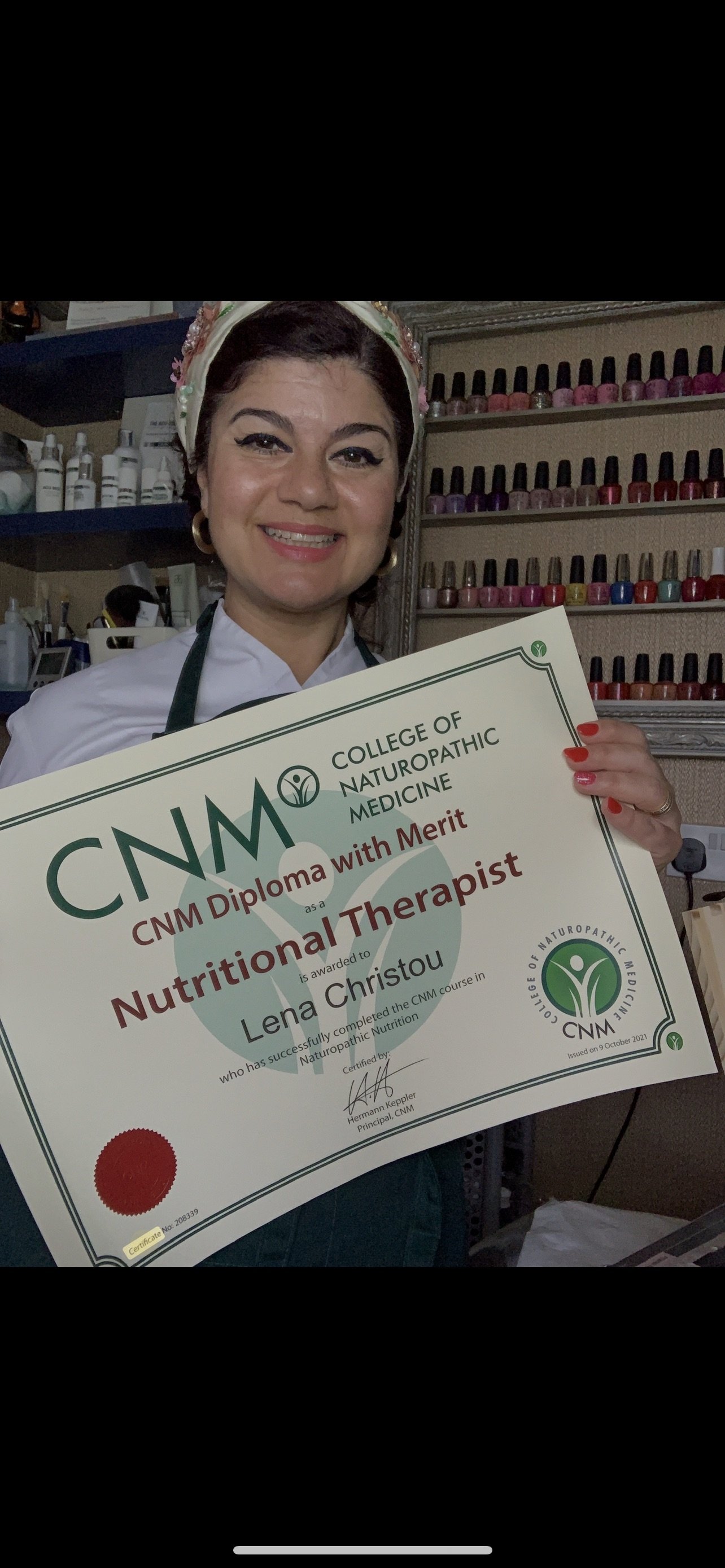 Woman holding a certificate from the College of Naturopathic Medicine, smiling, with nail polish and a headscarf, standing in front of shelves filled with colorful nail polish bottles.