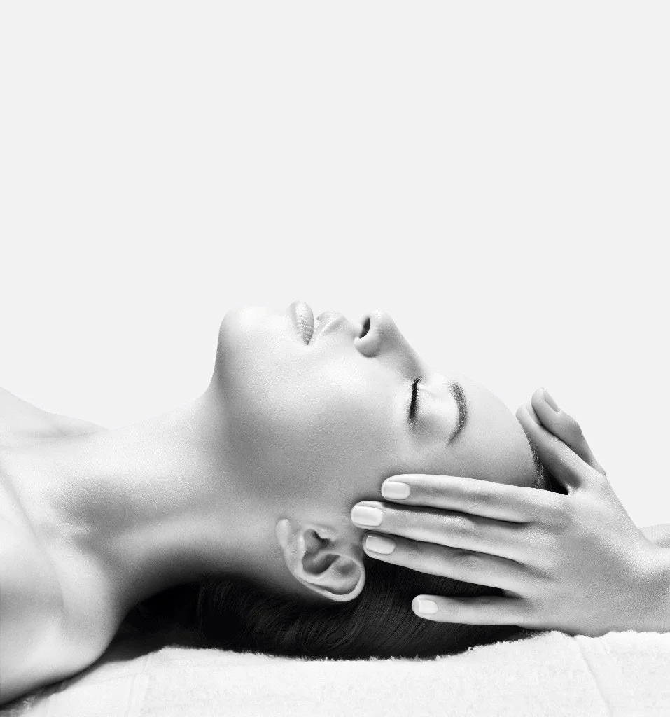A woman with closed eyes lying down while getting a massage or skin treatment on her face, with her hand gently touching her forehead.