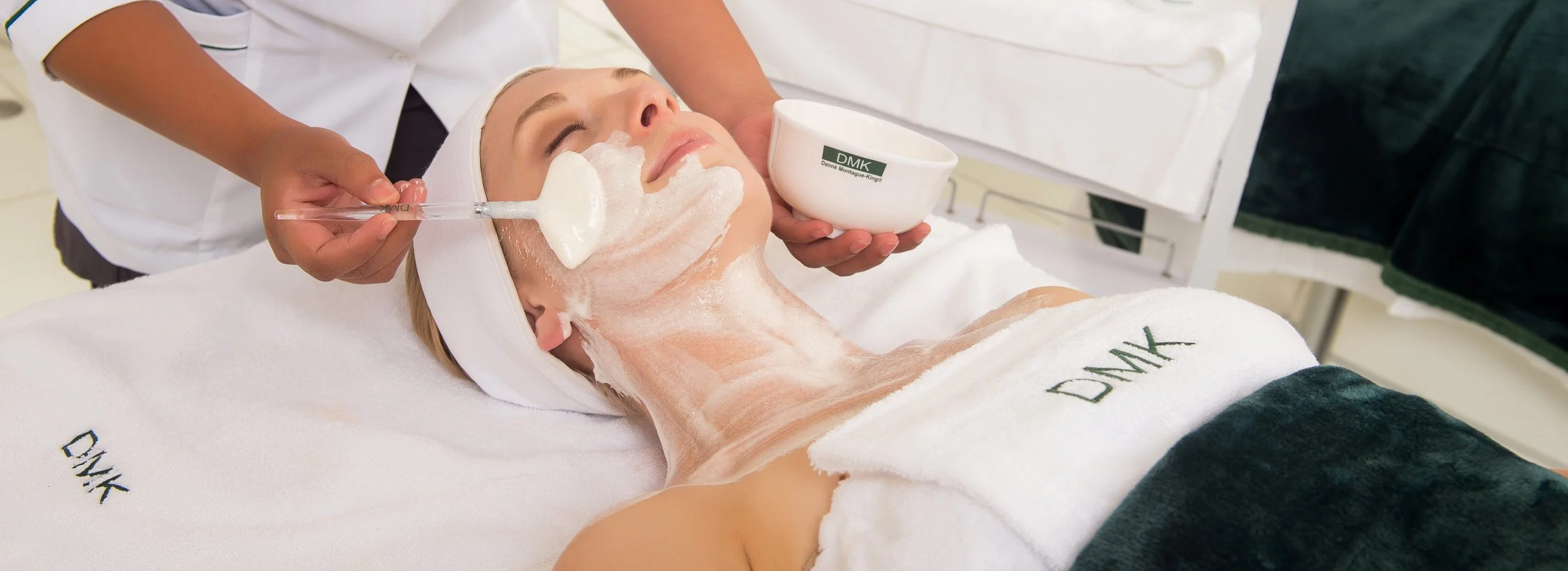 A woman getting a facial treatment at a spa, with a skincare product applied to her face and neck, while a skincare professional administers a facial mask or cream.