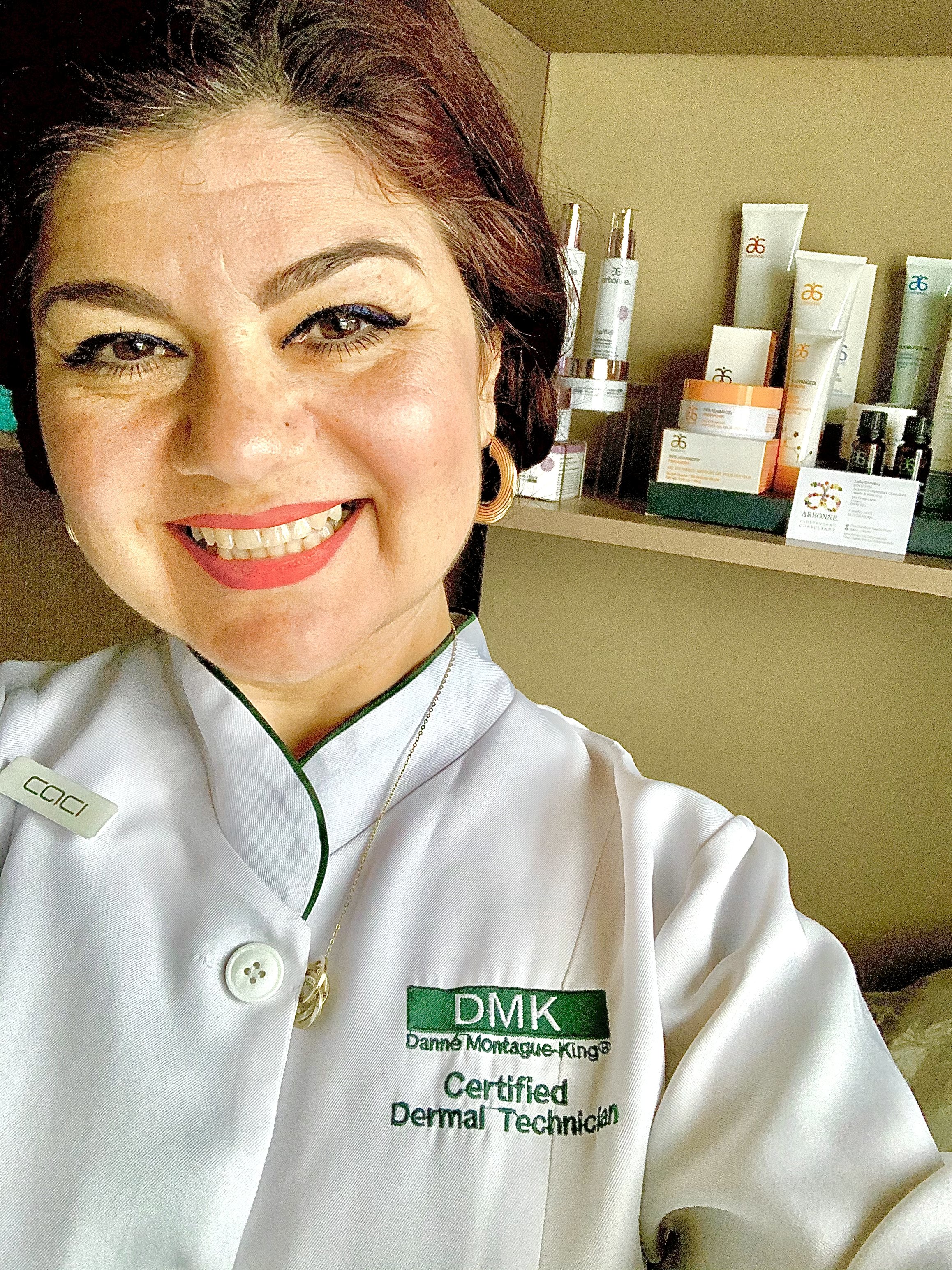 A woman smiling in a white uniform with a name tag and an embroidered patch that reads 'DMK', standing in front of shelves filled with skincare products and informational materials.