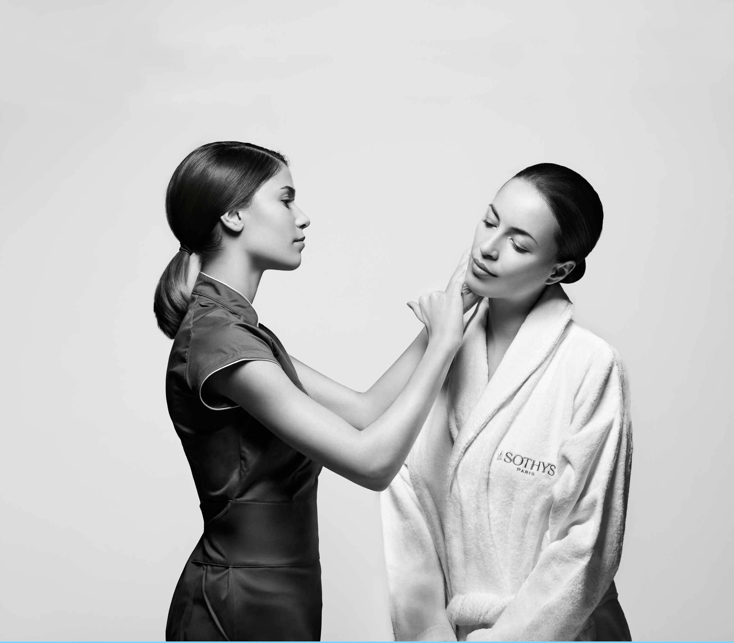 Two women with dark hair, closed eyes, and solemn expressions, one touching the other's face gently, set against a plain light background. The woman on the right is wearing a white robe with visible text 'SOTHYS PARIS,' and the woman on the left is in a dark top.