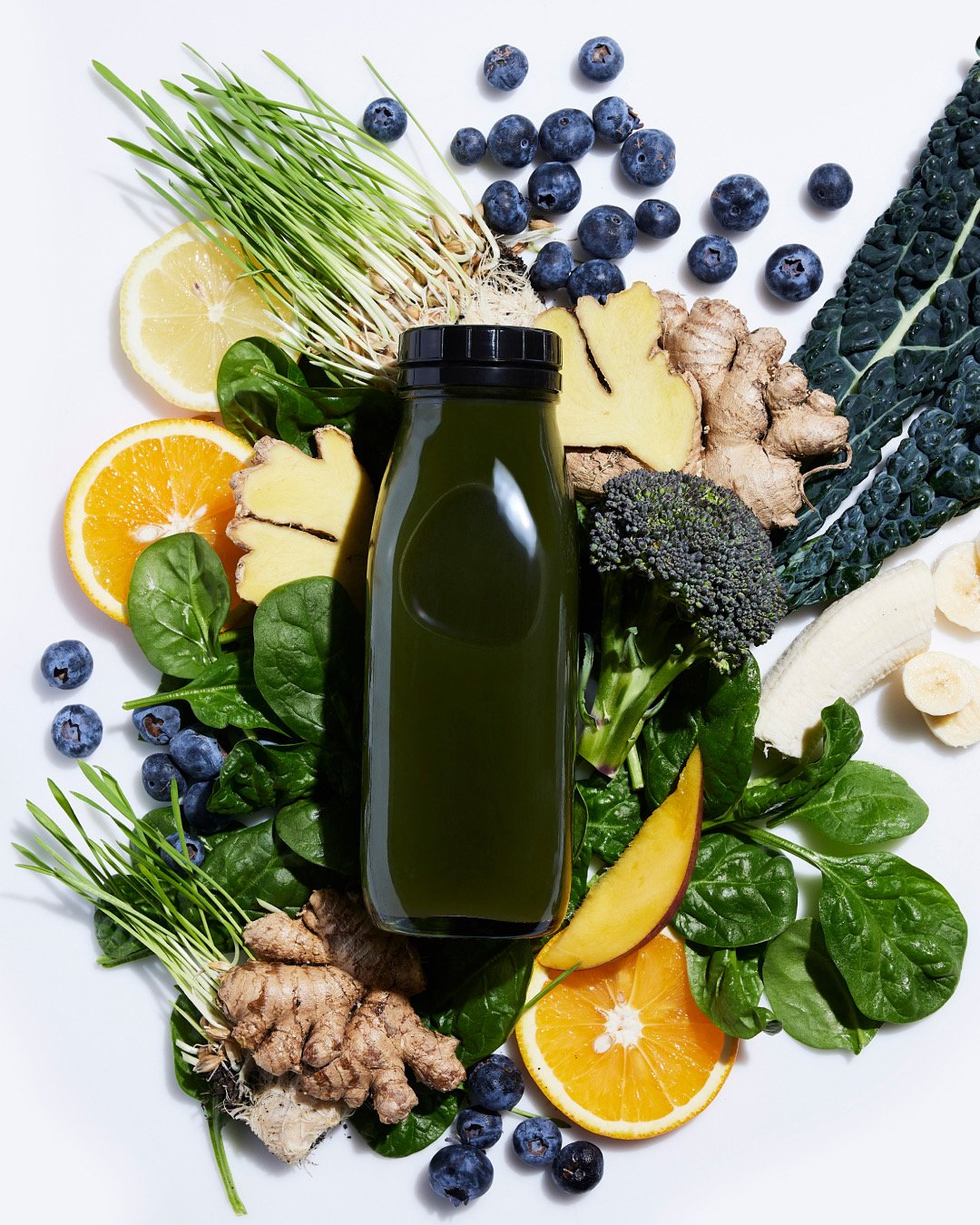A bottle of green juice surrounded by blueberries, lemon slices, orange slices, ginger, kale, spinach, broccoli, banana slices, and ginger root on a white surface.