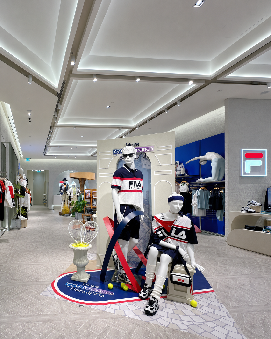 Clothing store display with mannequins wearing sporty outfits, tennis rackets with tennis balls, and sneakers, featuring a backdrop with sports-themed decor and the branding 'FILA'.
