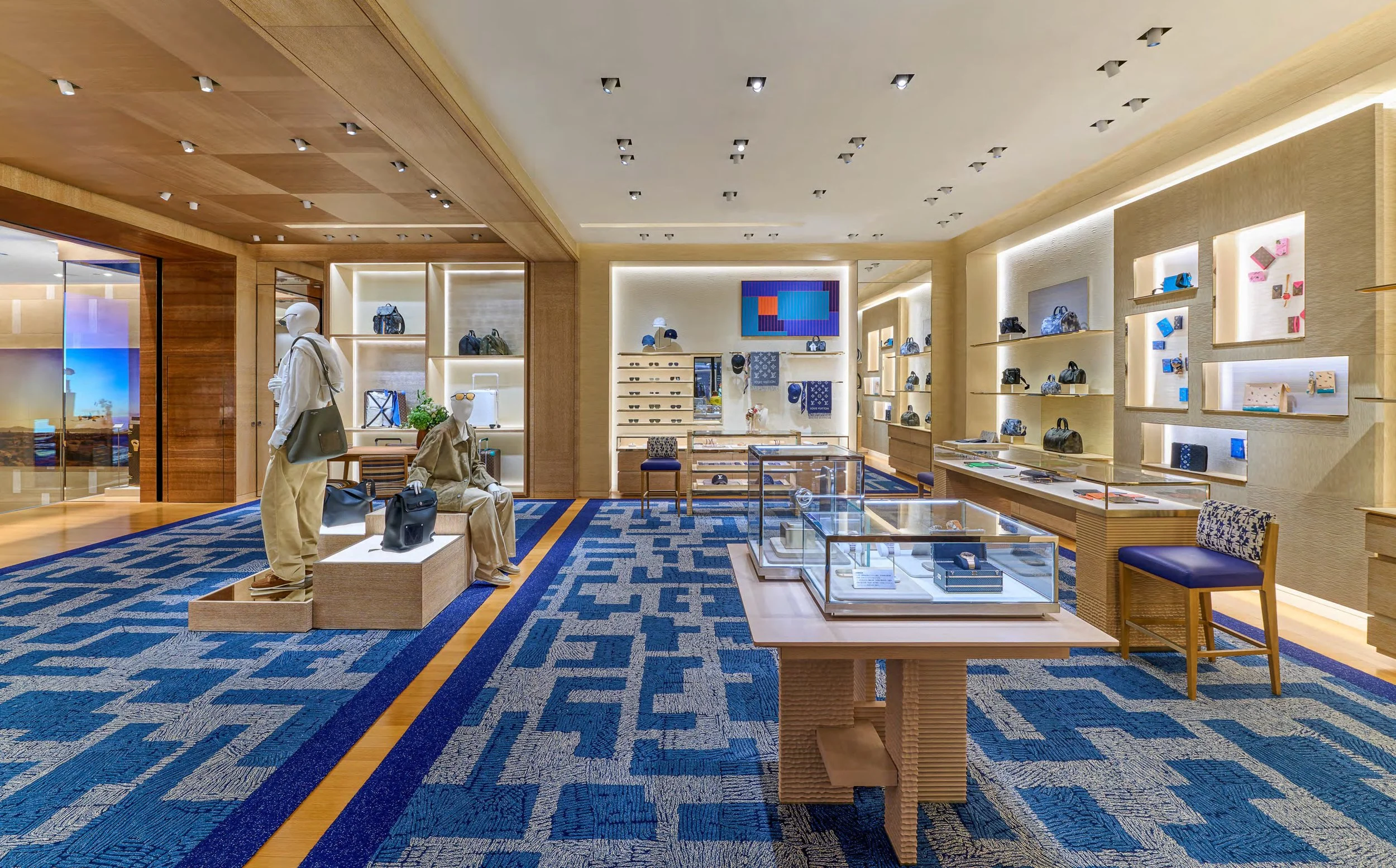 Interior of a retail store with displays of handbags, sunglasses, and accessories, featuring wood shelving, glass display cases, and mannequins, with blue patterned carpet and recessed ceiling lights.