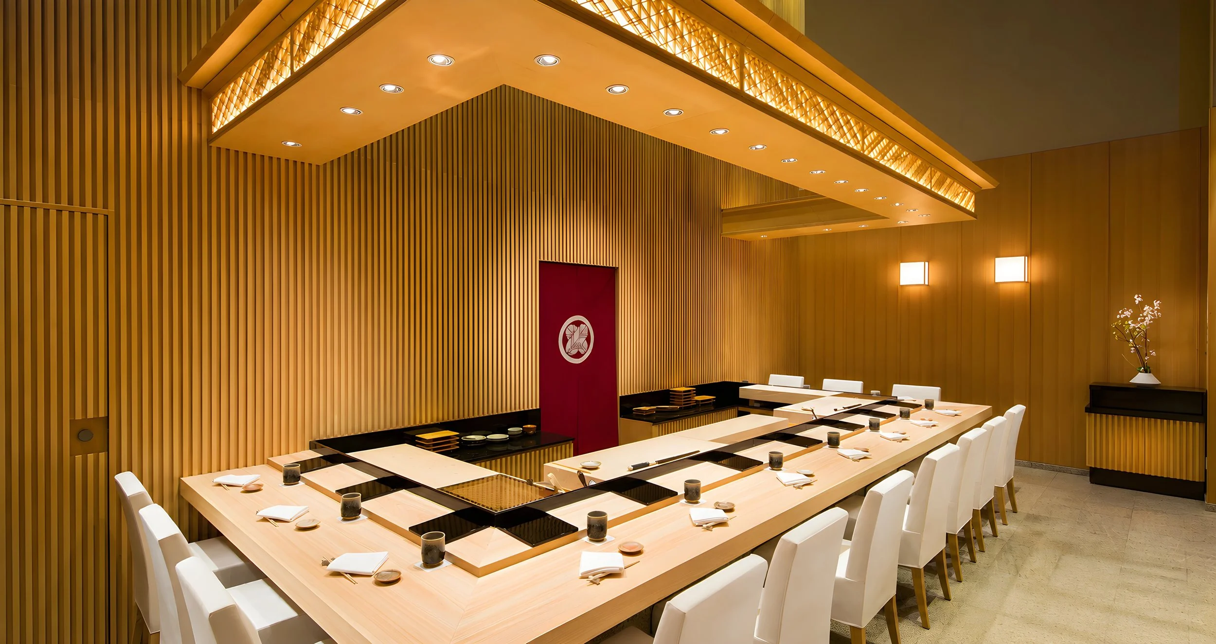 A modern Japanese restaurant interior with a rectangular wooden dining table set with plates and cups, surrounded by white chairs, wooden slat walls, and warm lighting, featuring a red door with a white emblem and a potted orchid on a black side table.