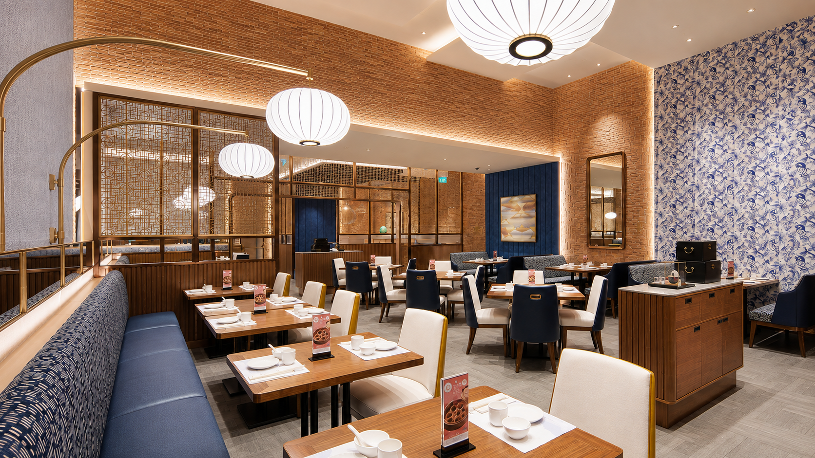 A modern restaurant dining area with tables set with white bowls and utensils, surrounded by a mix of blue, white, and beige chairs, featuring a decorative blue floral wallpaper on one wall, warm lighting, and stylish hanging light fixtures.