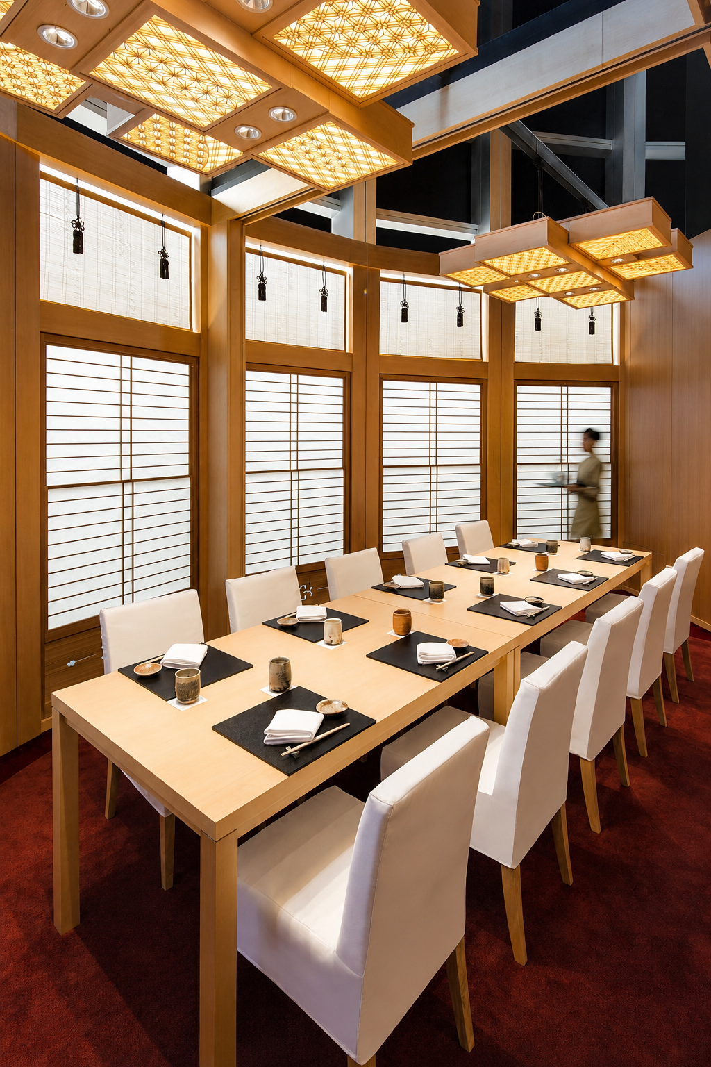Elegant dining room with a long wooden table set with black placemats, white napkins, and small cups, surrounded by white upholstered chairs. The room features Japanese-style shoji screens, warm wooden accents, and large decorative ceiling lights with a geometric design. A woman in traditional attire is seen walking in the background.