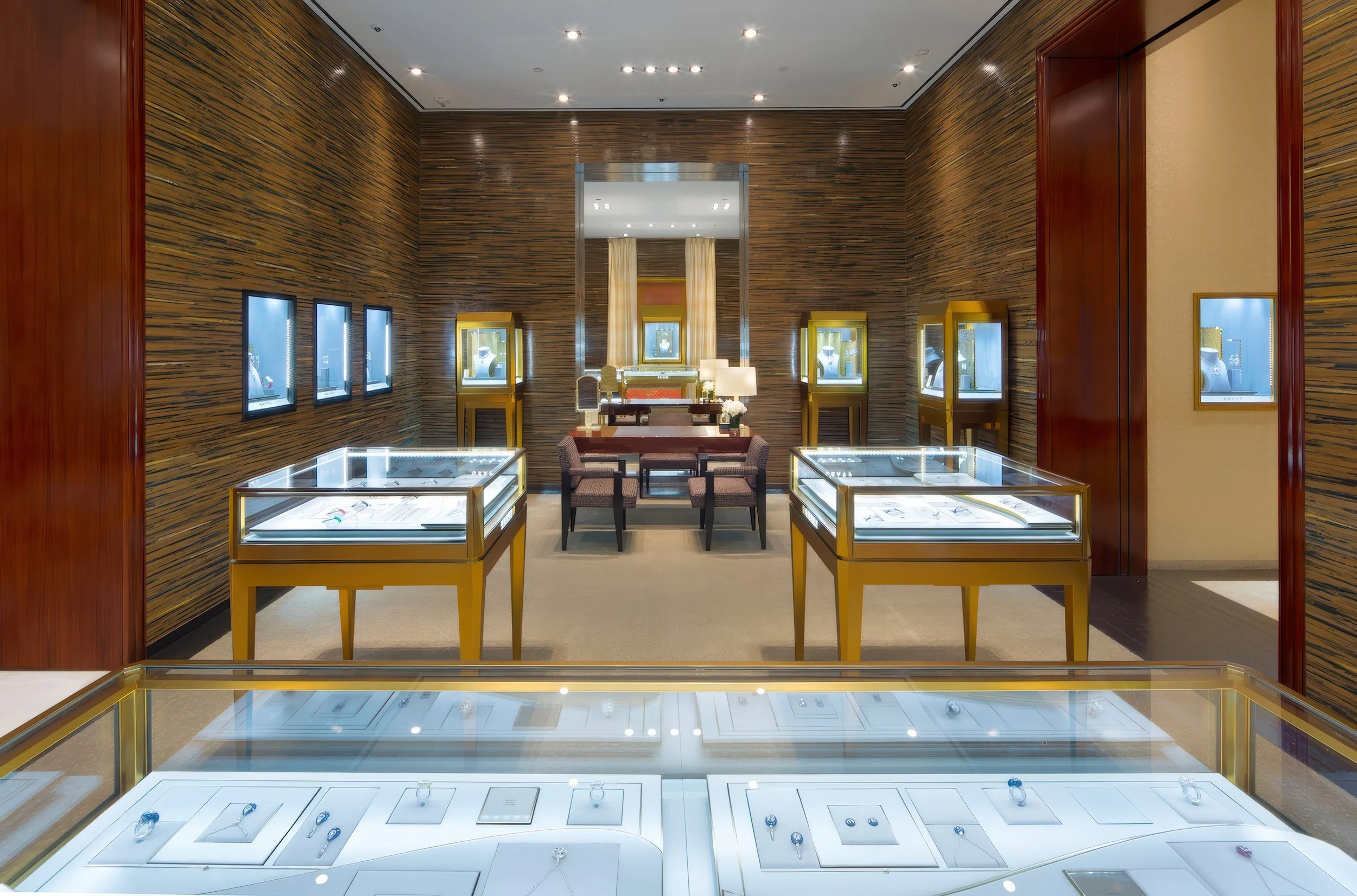 View of a jewelry store display area with glass cases and illuminated jewelry pieces, wooden walls, and a seating area with chairs and lamps.