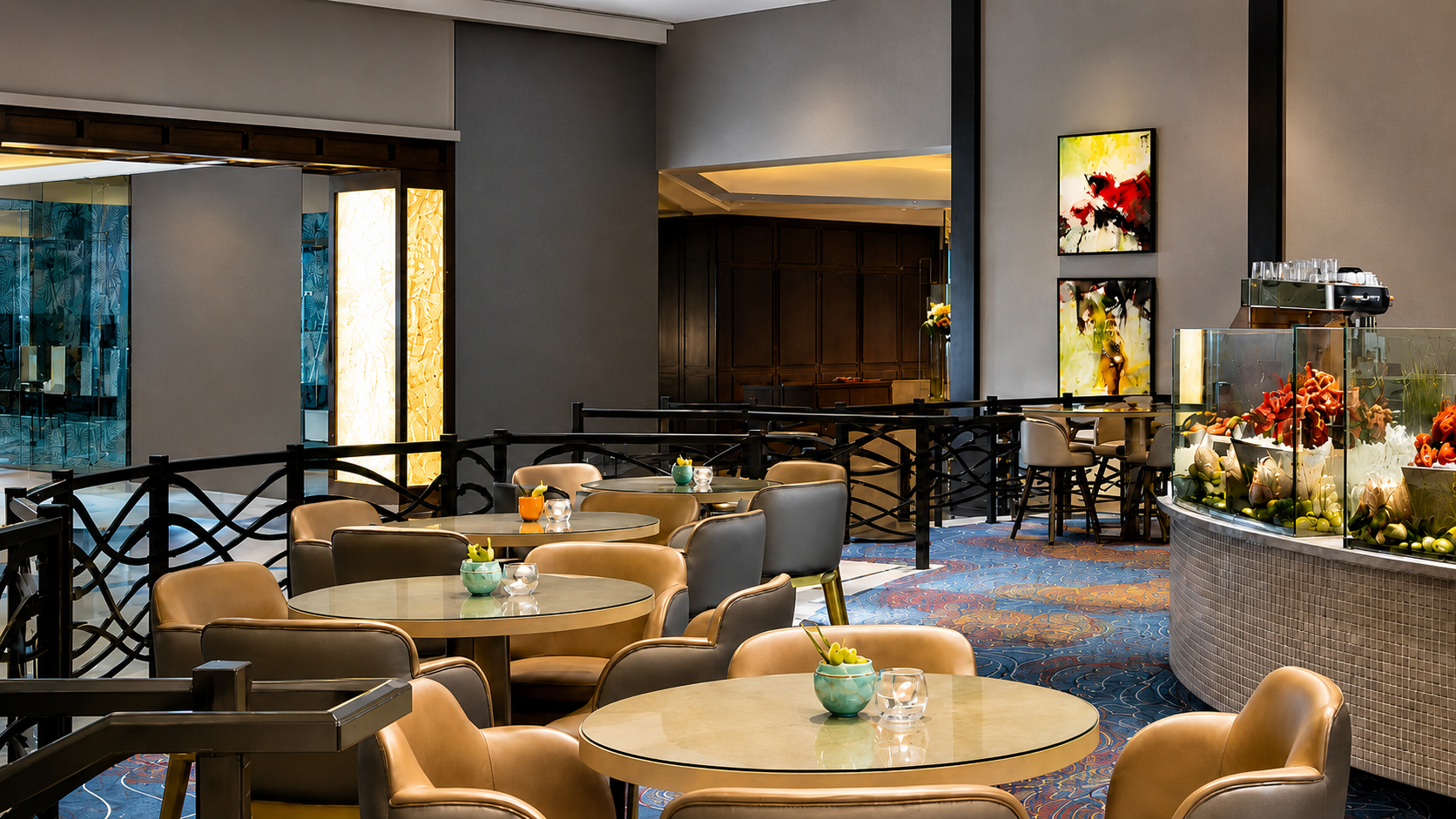 Elegant hotel dining area with round tables, beige chairs, decorative floral arrangements, abstract artwork on walls, and a curved glass display case with flowers.