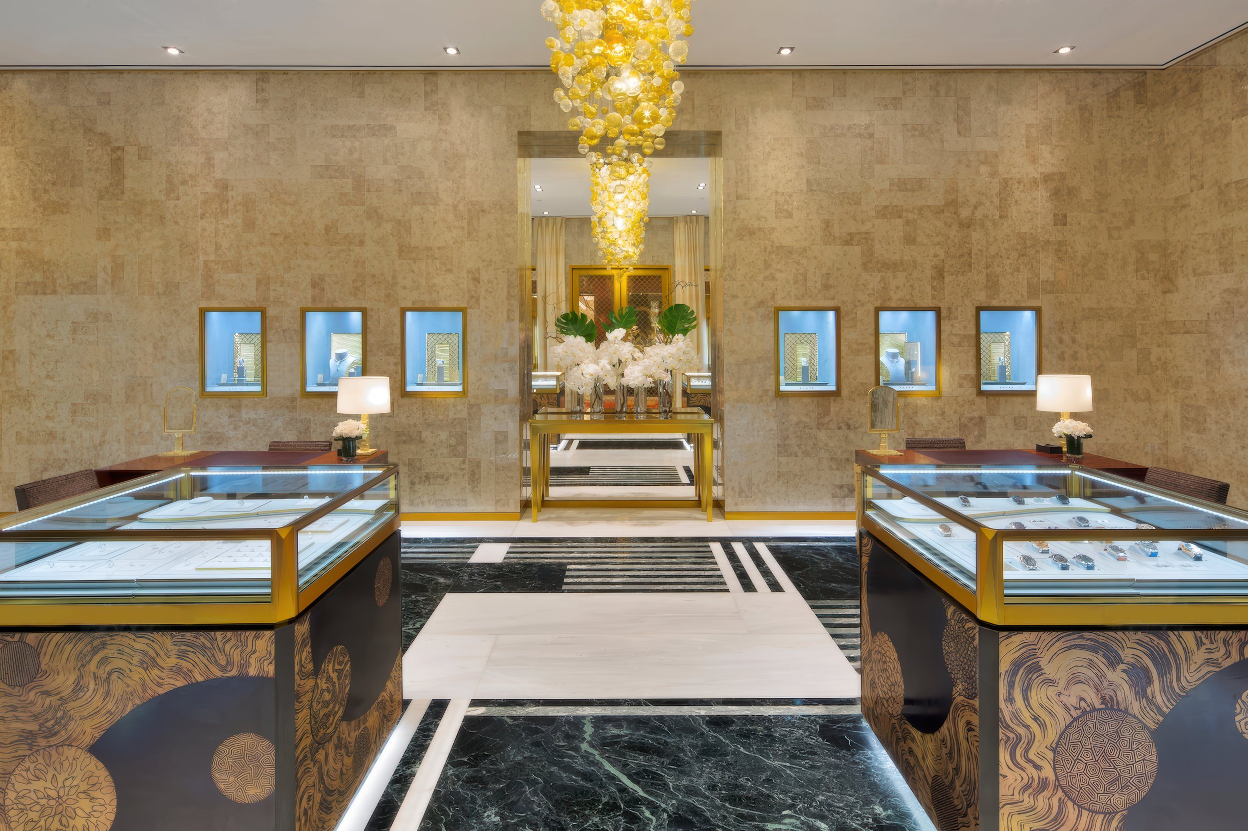 Inside a jewelry store with gold accents, display cases with jewelry, a large floral arrangement with white flowers and large green leaves, and a decorative chandelier hanging from the ceiling.