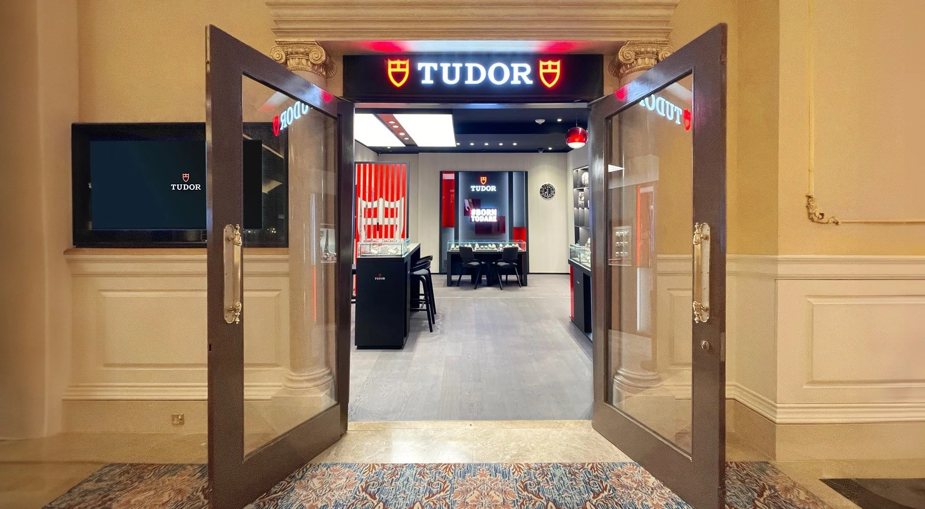 Entrance to a Tudor jewelry store with open glass doors, displaying the store's logo and interior lighting and jewelry displays inside.