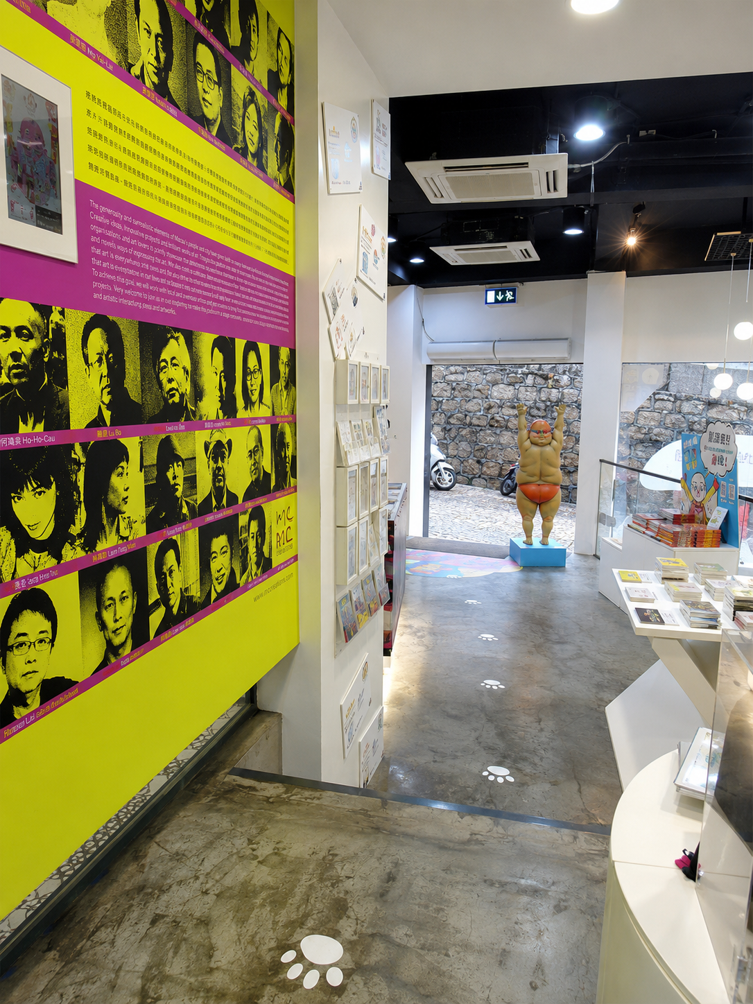 Inside a bookstore or exhibit space featuring a large yellow panel with black and white portraits of various people, a colorful sculpture of a sumo wrestler at the entrance, and shelves with books and artwork.