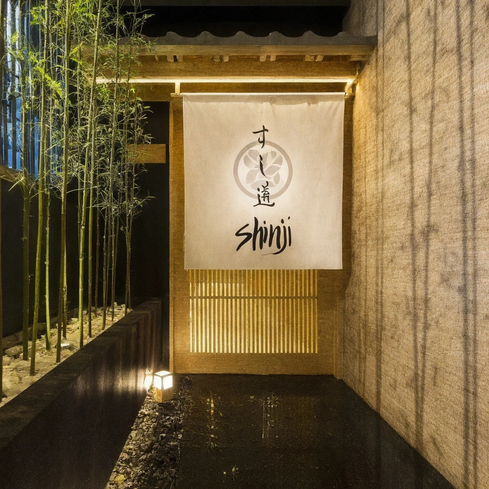 Nighttime view of a vertical fabric sign with Japanese characters and the word 'shinji,' illuminated from below, situated next to bamboo plants on the left and a beige textured wall on the right, with soft lighting creating shadows.