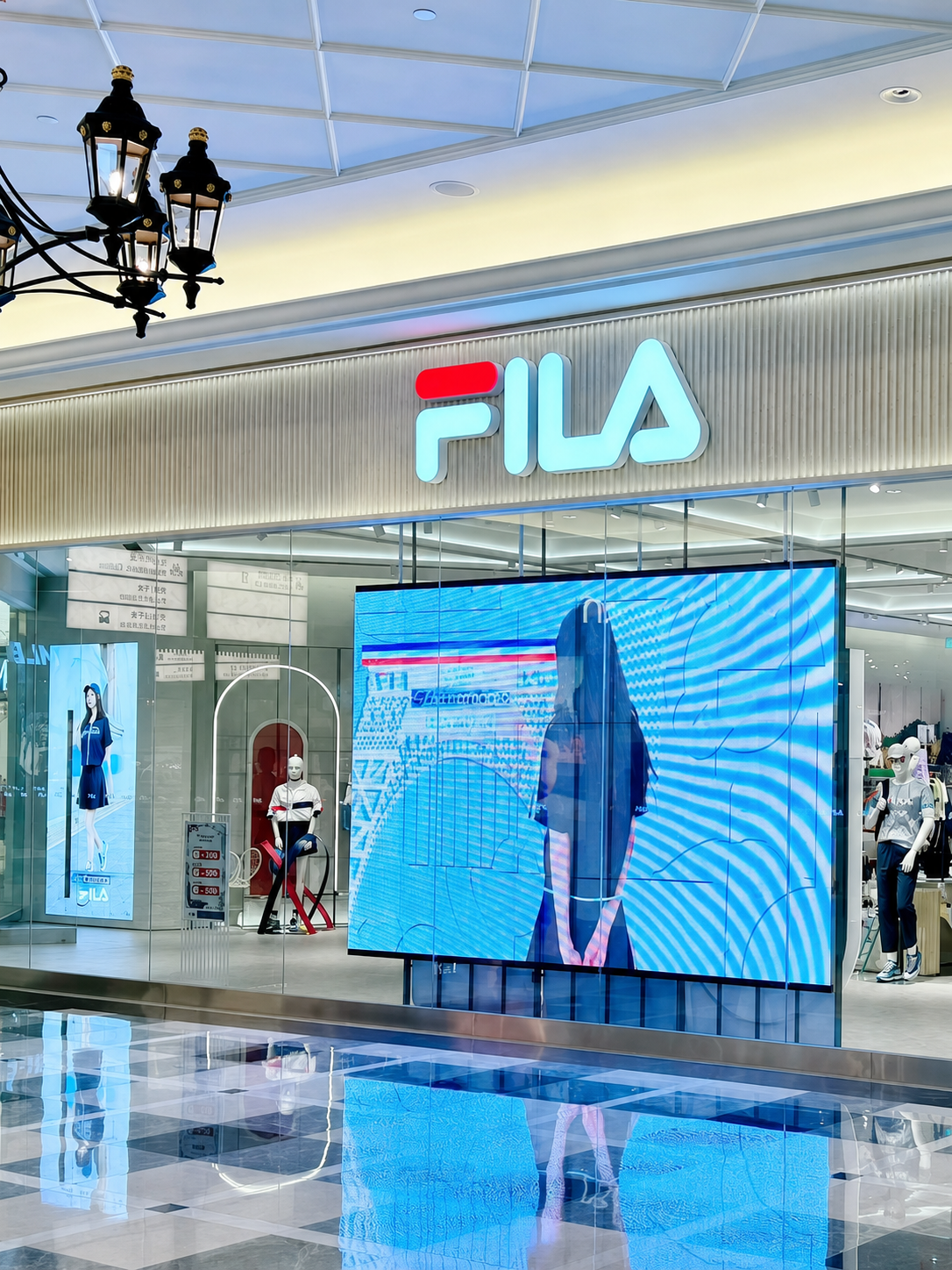 Exterior of a FILA retail store in a shopping mall with mannequins and a digital screen displaying fashion advertisements.