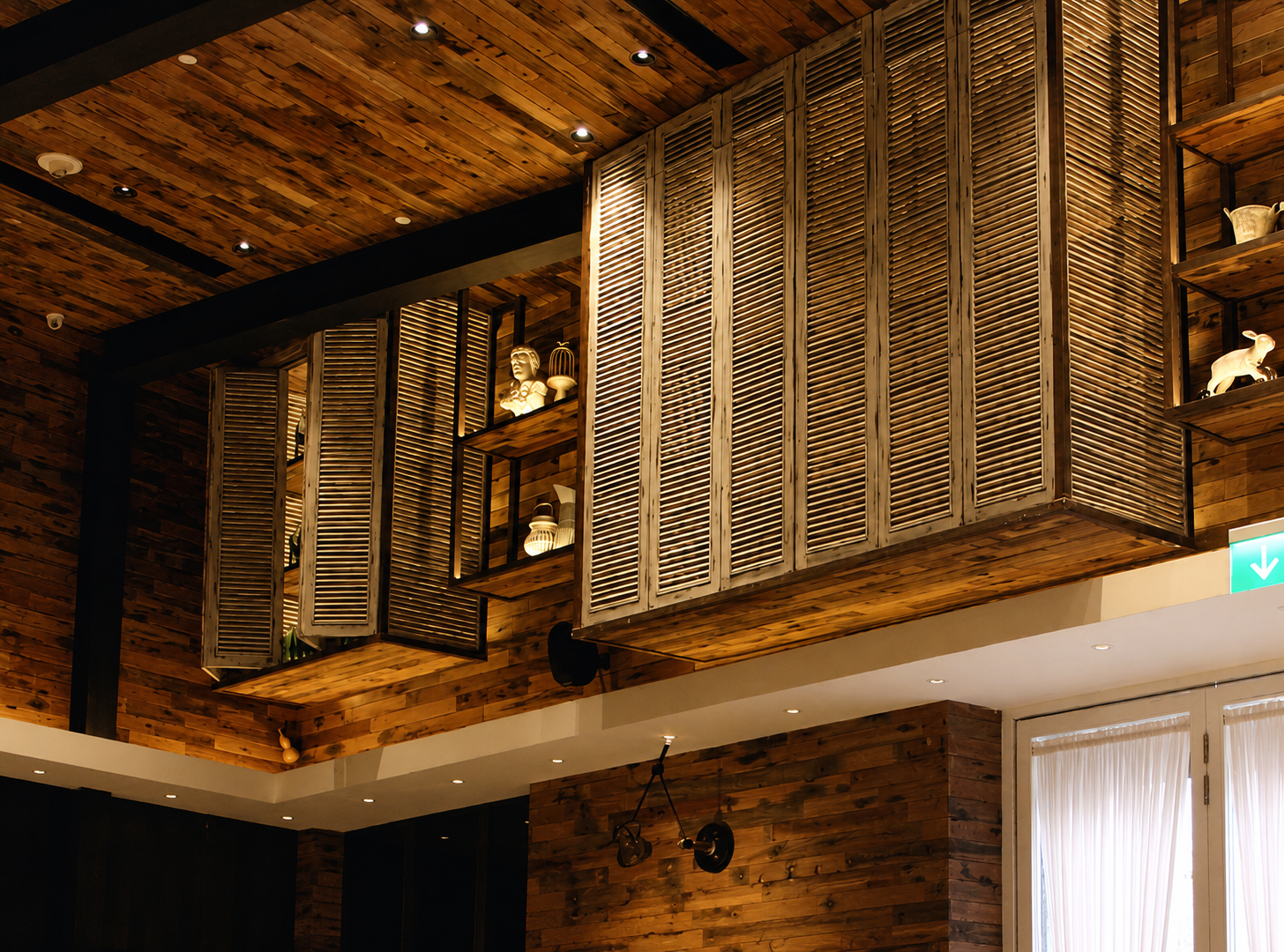Interior of a room with a wooden ceiling and walls, featuring decorative wooden shutters and shelves displaying sculptures and lit lamps.
